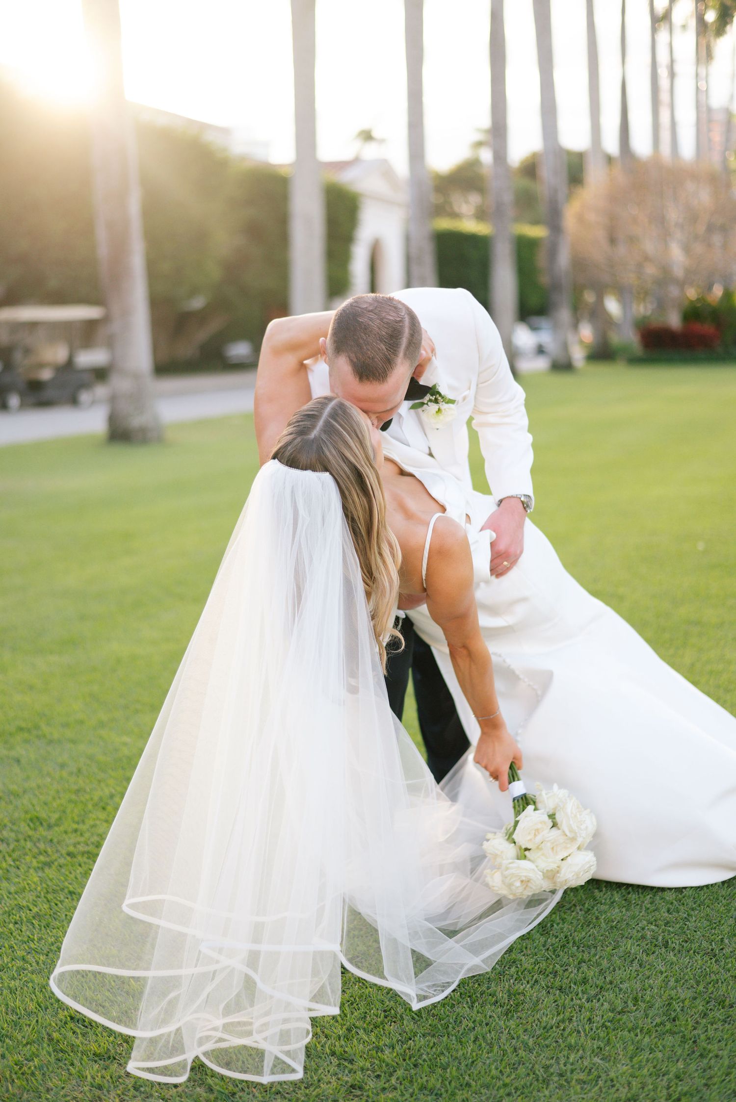 Wedding couple embracing on a sunlit lawn with flowing white veil.