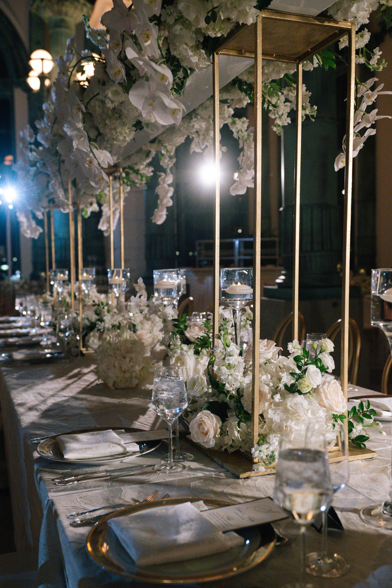 Tall gold geometric centerpieces with white orchids and florals adorn elegant wedding reception tables.