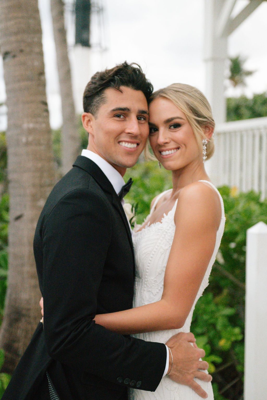 Newlyweds embrace on white porch with tropical greenery in background.