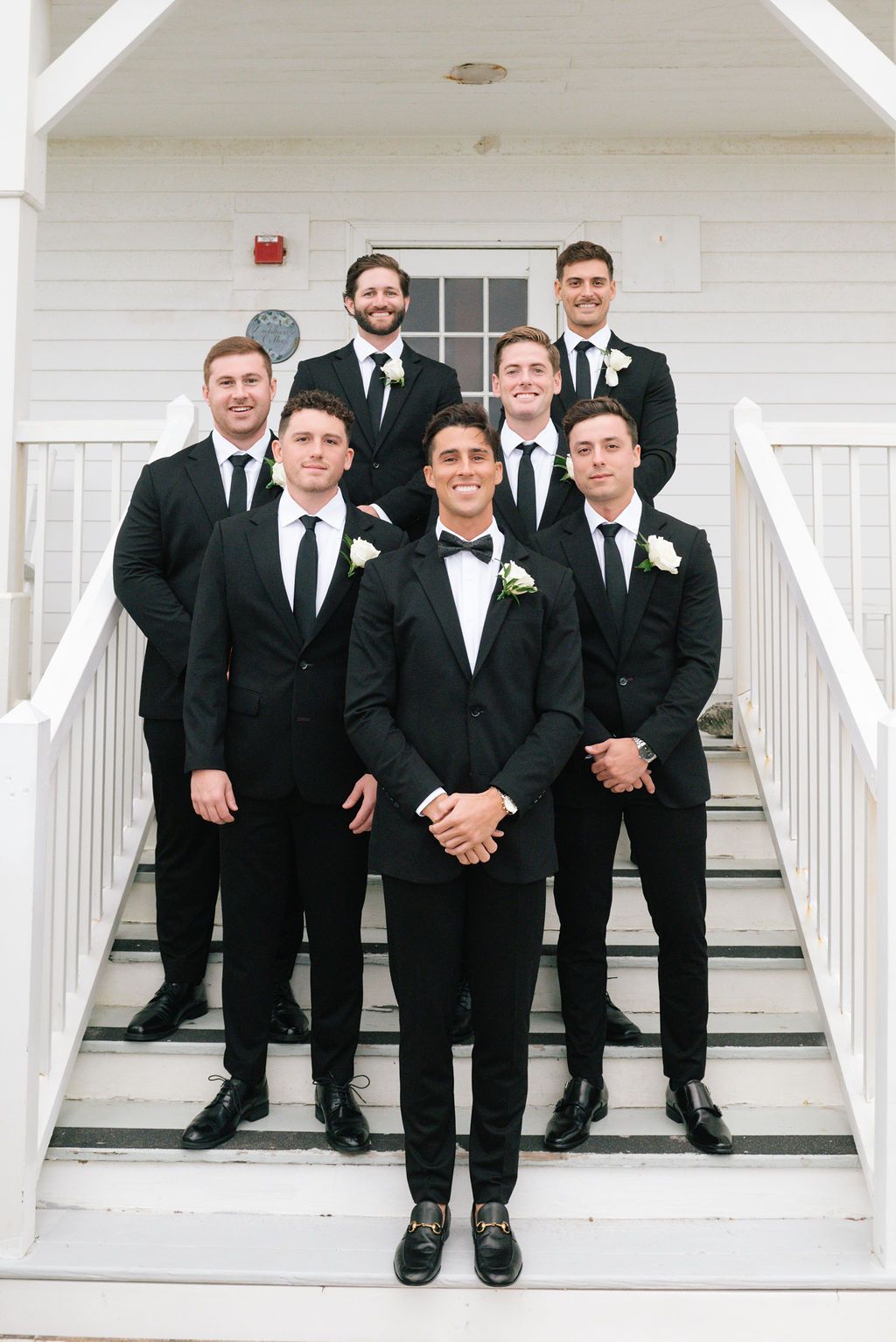 Groomsmen in black tuxedos pose on white porch steps of a building.