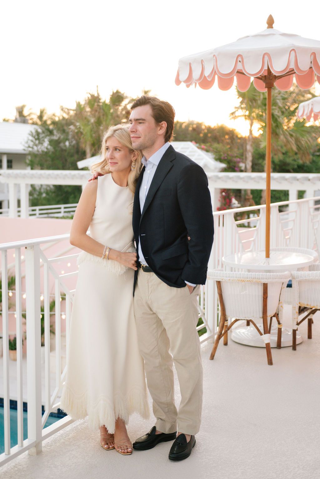 Couple embracing on balcony with coral umbrella and sunset backdrop.