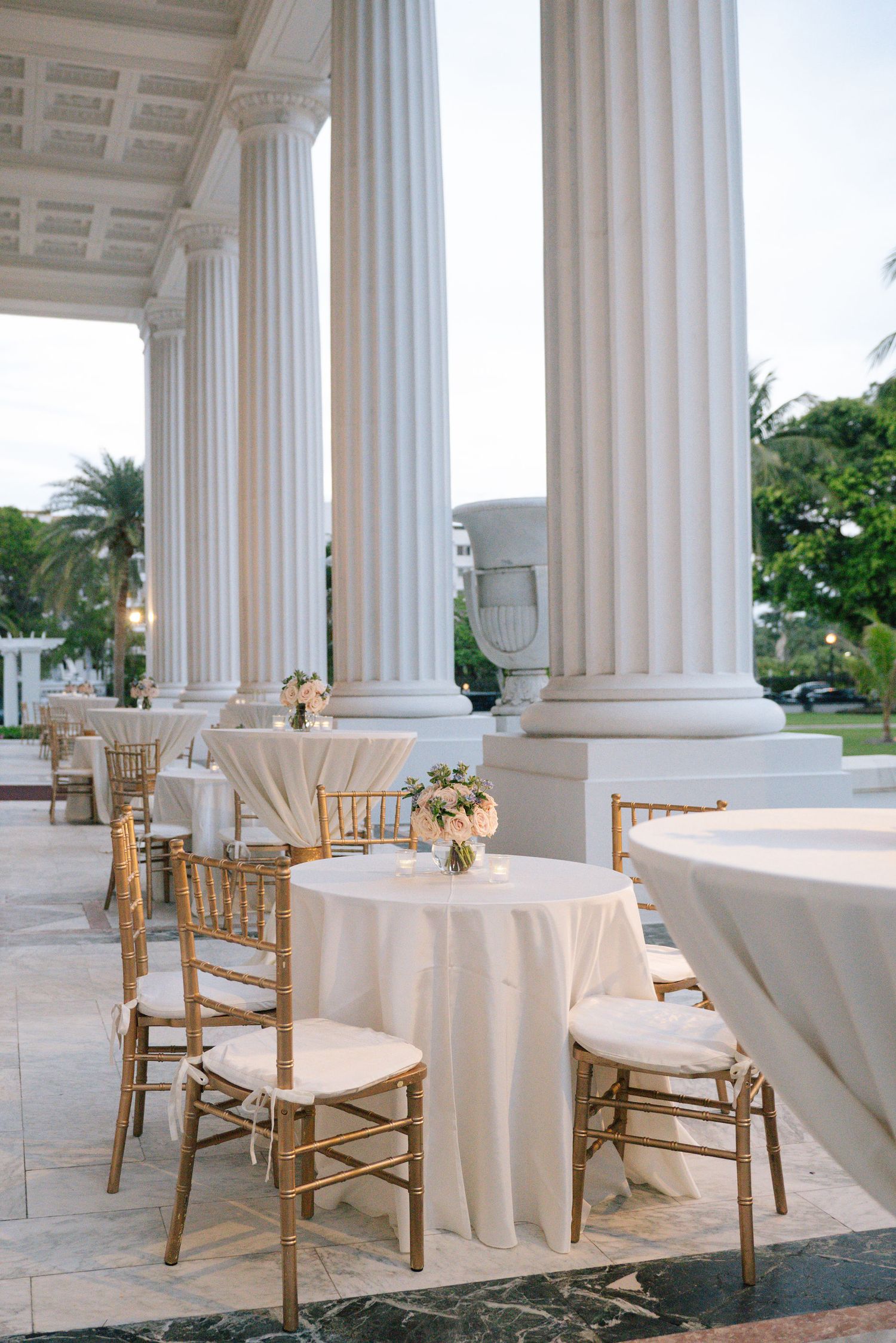 Elegant wedding cocktail setup with white linens and gold chiavari chairs between classic white columns on a terrace.