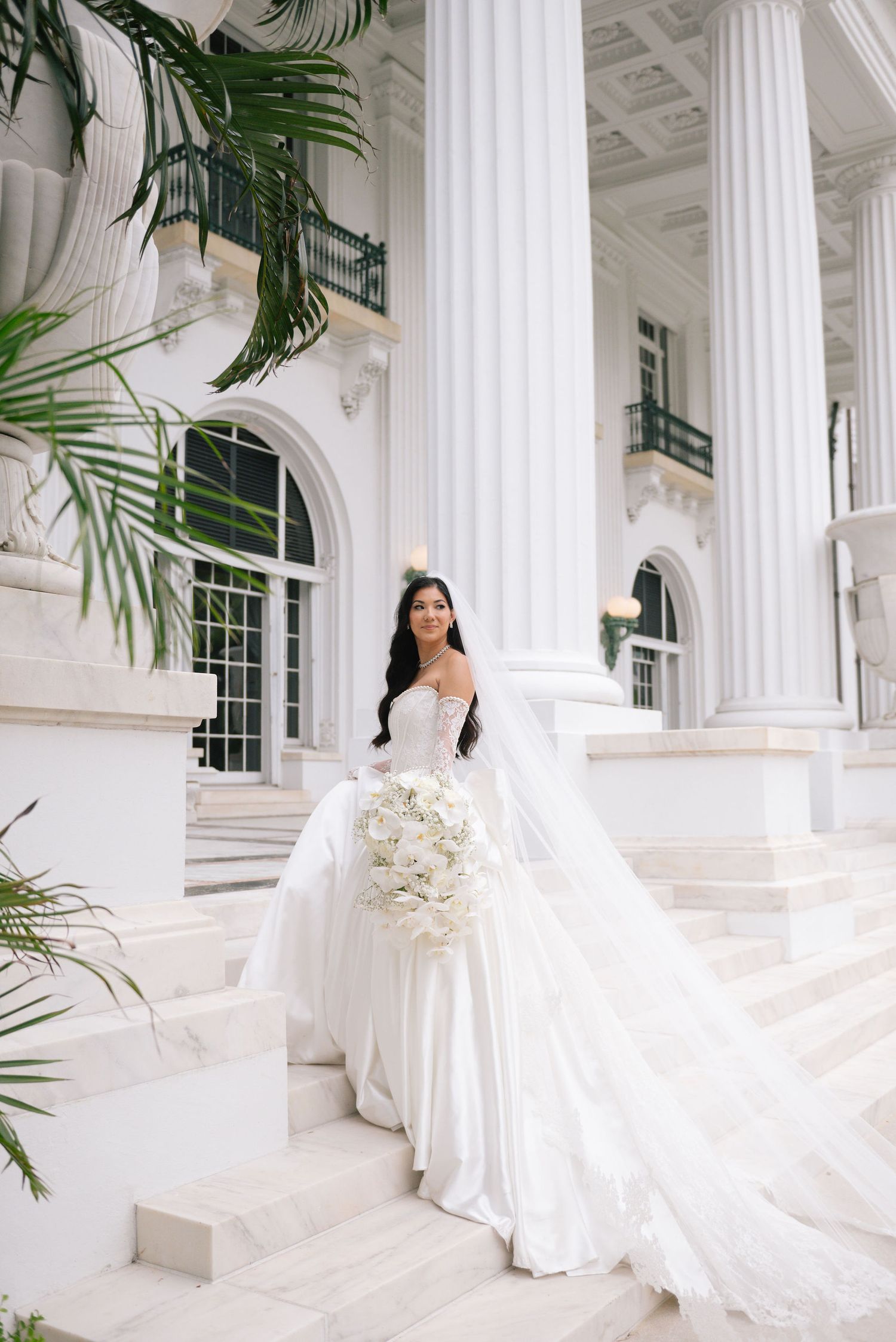 A bride in an elegant white gown poses among classical white columns with tropical greenery.