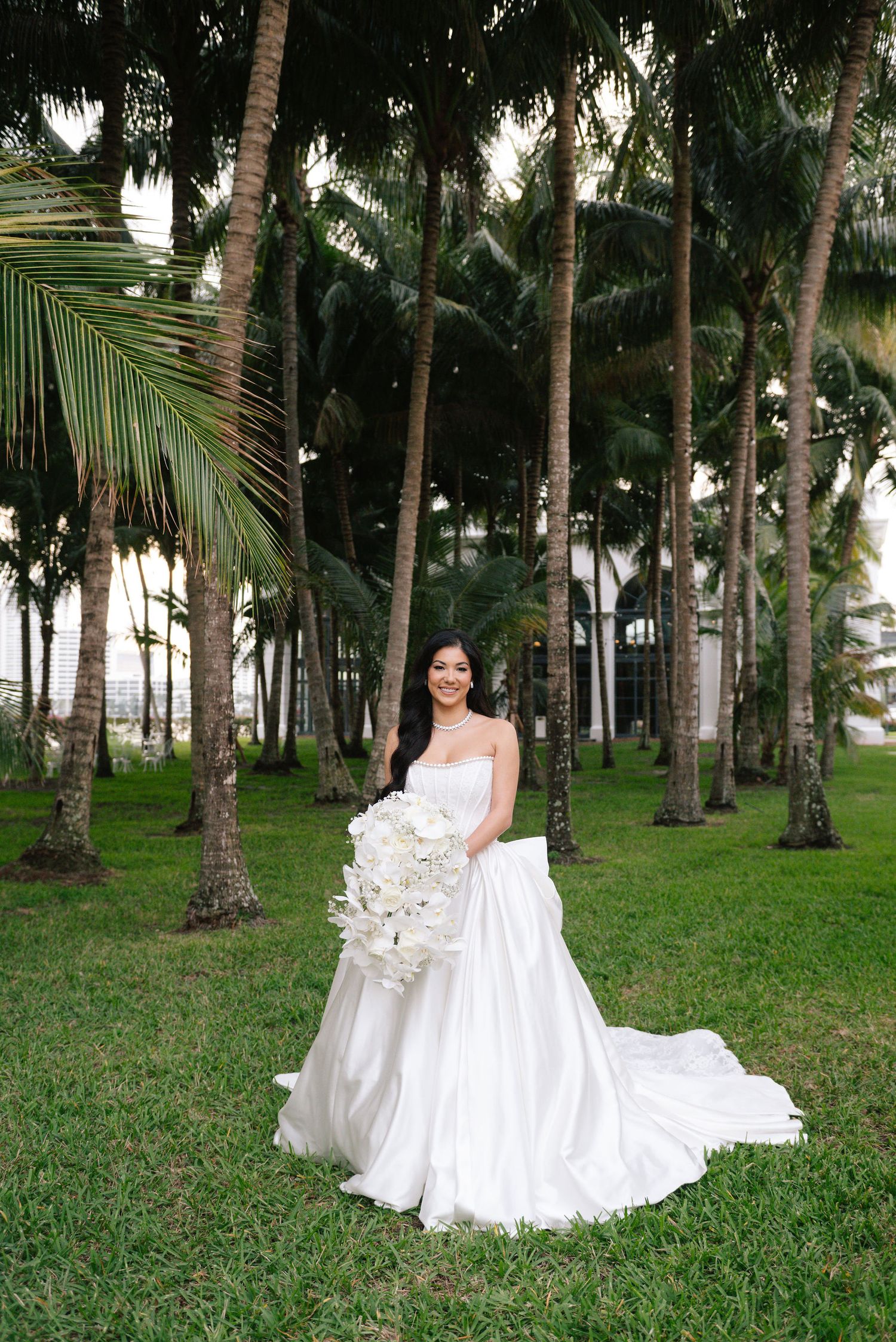 A bride in a voluminous white gown poses gracefully among palm trees in a tropical garden setting.