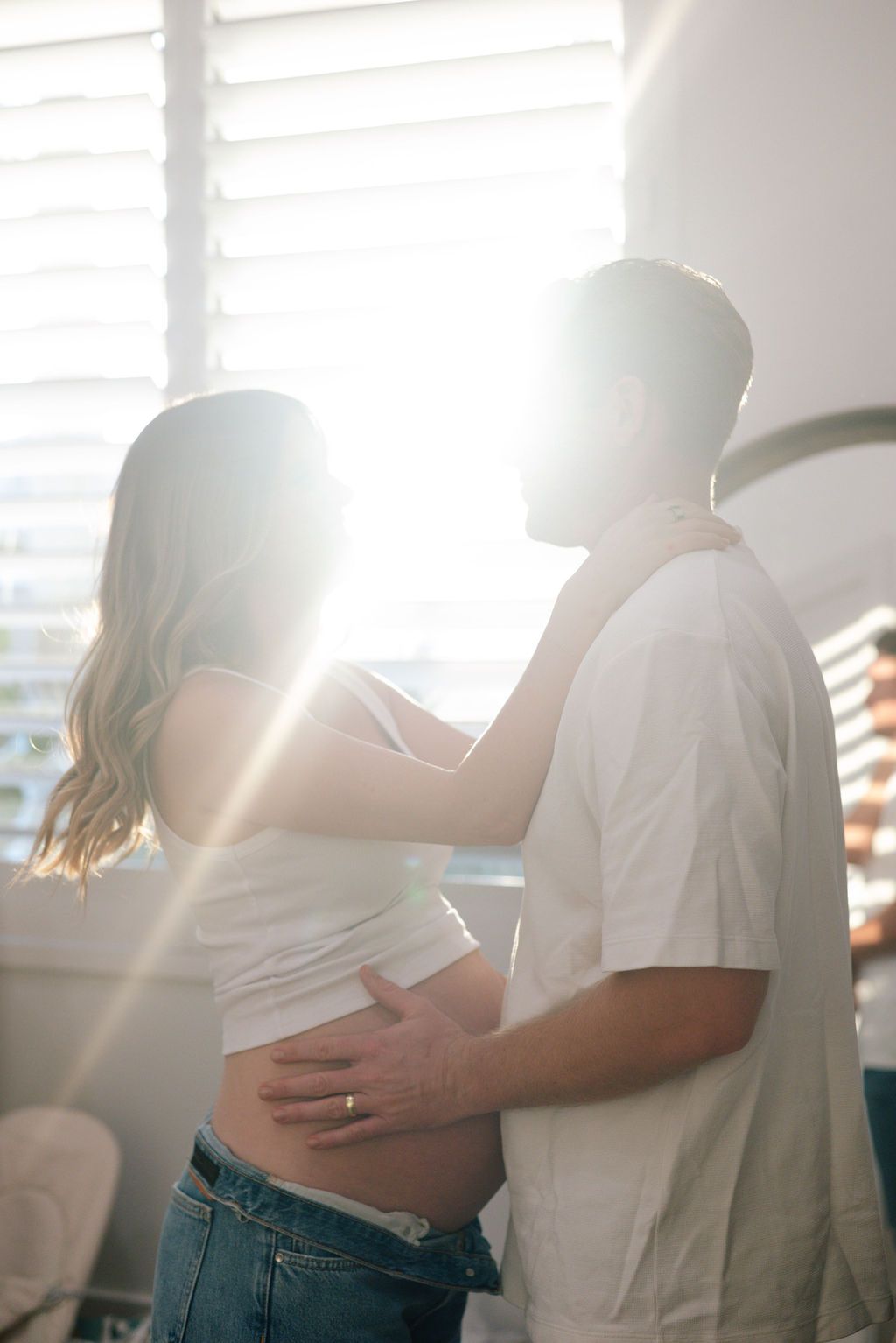 Sunlit intimate moment between couple in white tops and jeans by window.