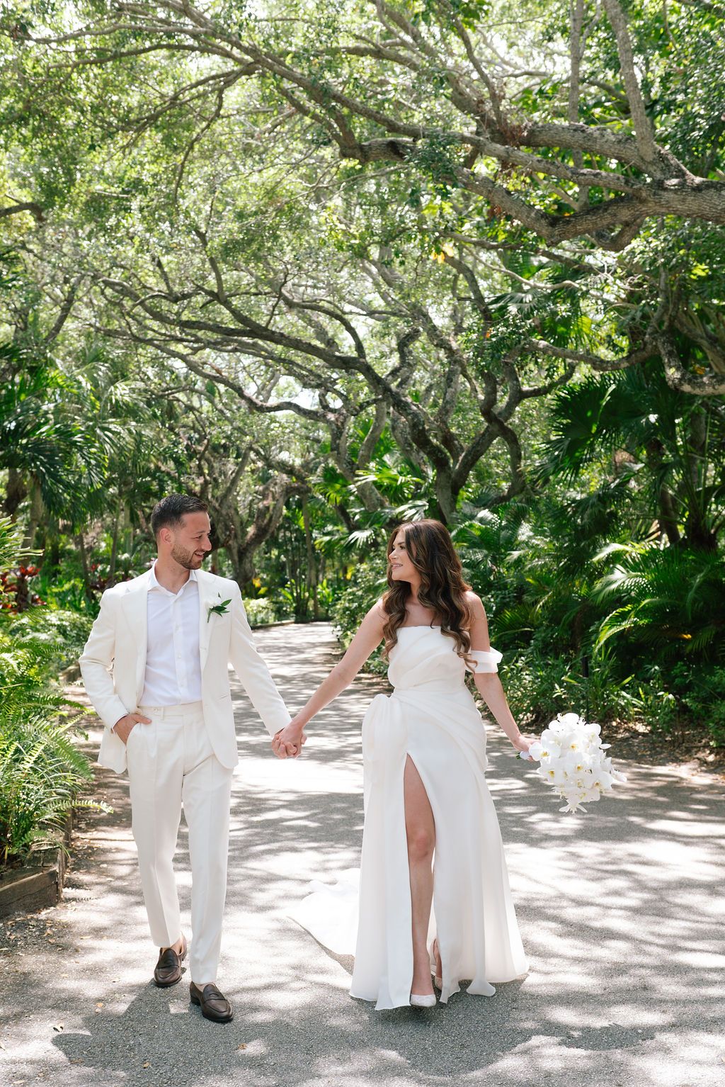 Wedding couple in white sharing playful moments on a sun-dappled garden path.