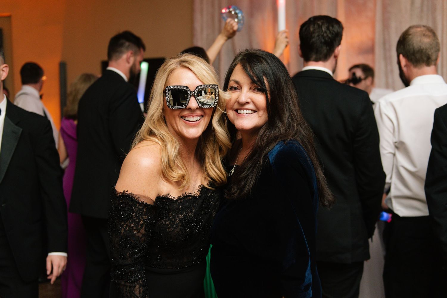 Two women in evening wear smile together at a formal event with other guests visible in the background.