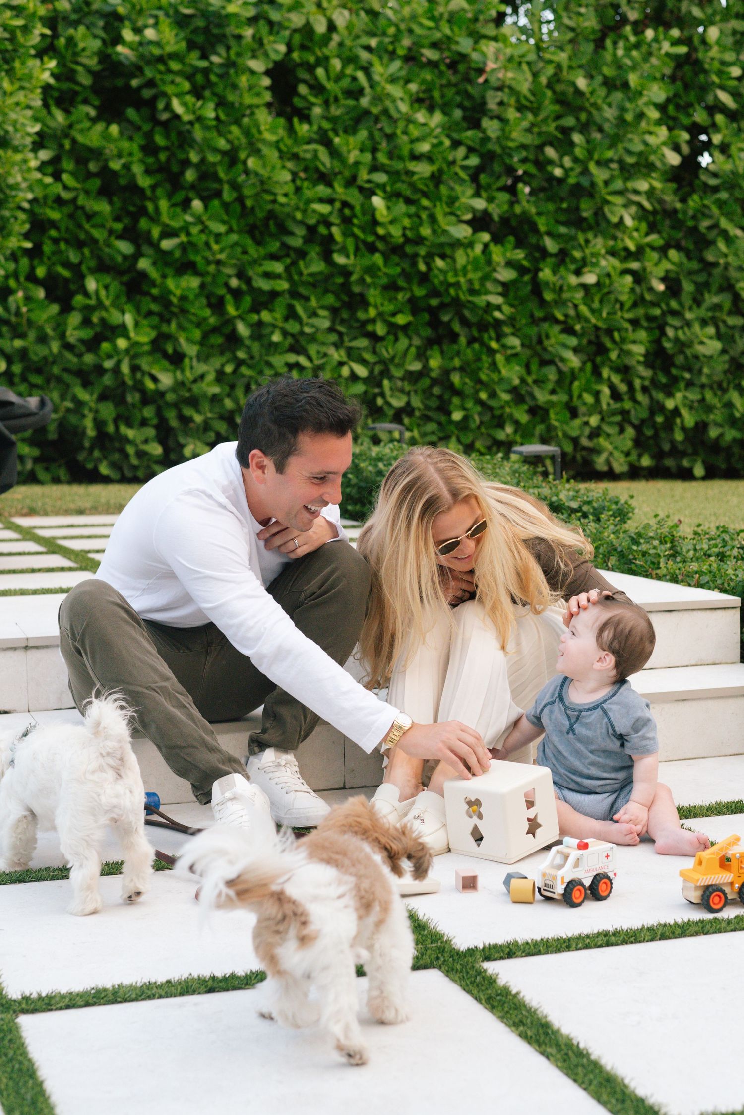 Family relaxing on white outdoor patio with small dog and toys in lush garden setting.