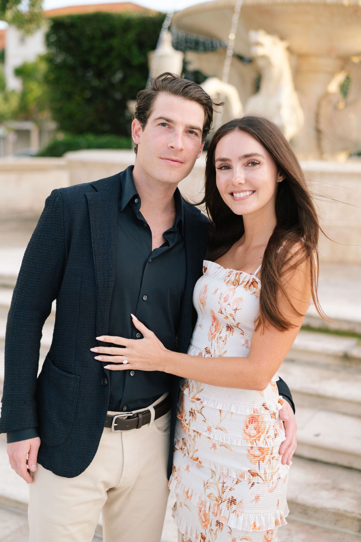 A couple poses together outdoors, with her wearing a floral dress and him in a dark blazer and khakis.