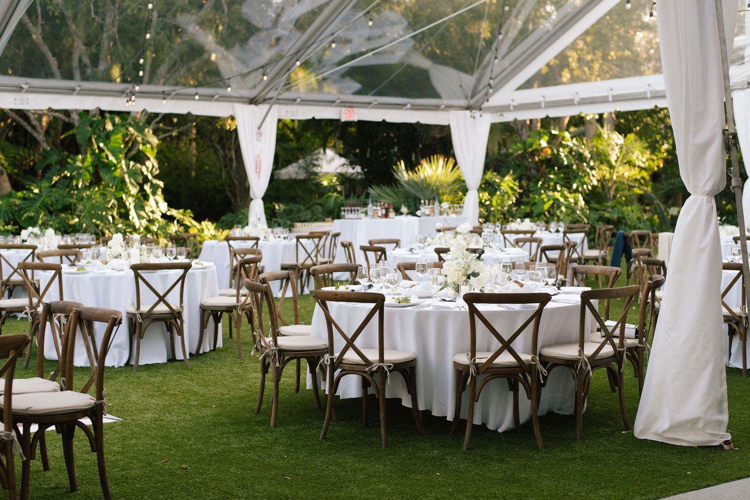 An elegant outdoor wedding reception setup under a white tent with cross-back chairs and round tables on grass.