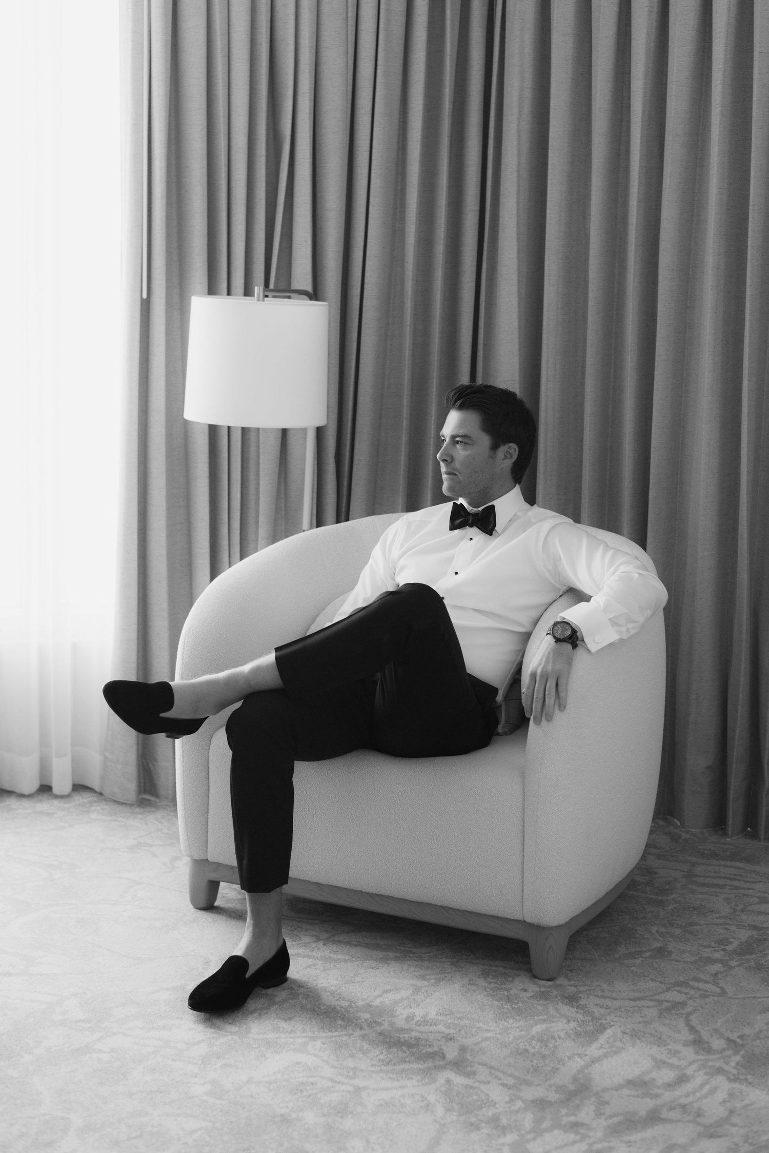 Black and white photo of a person in formal attire lounging on a white armchair against curtains with a lamp in background.