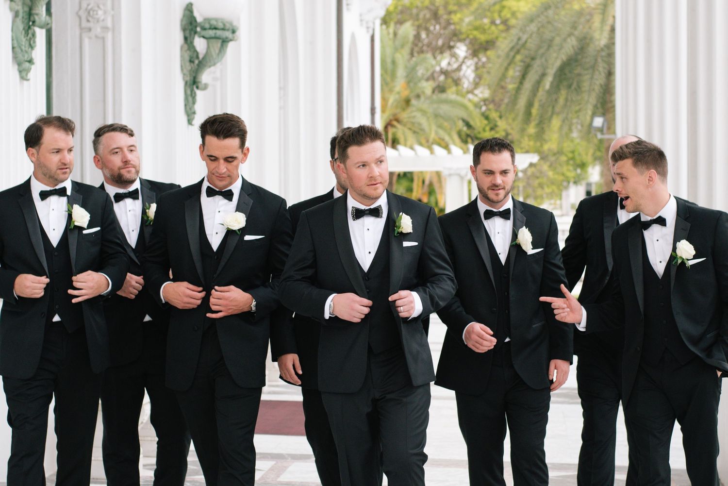 Seven groomsmen in black tuxedos with bow ties and white boutonnieres walk together at an elegant outdoor wedding venue.