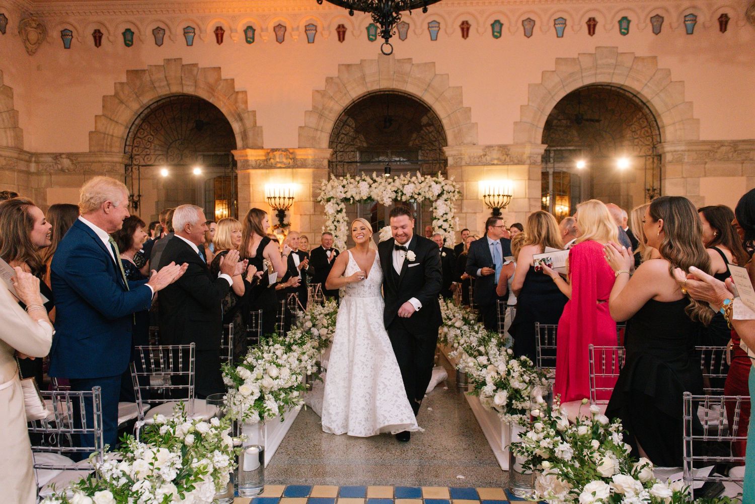 Bride and groom walking down aisle at elegant indoor ceremony with white floral decor.