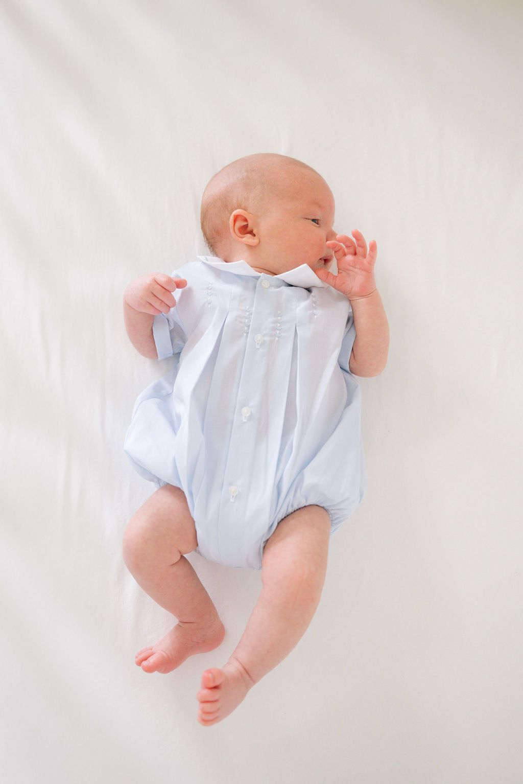 Baby in pale blue romper stretches against white backdrop in natural lighting.
