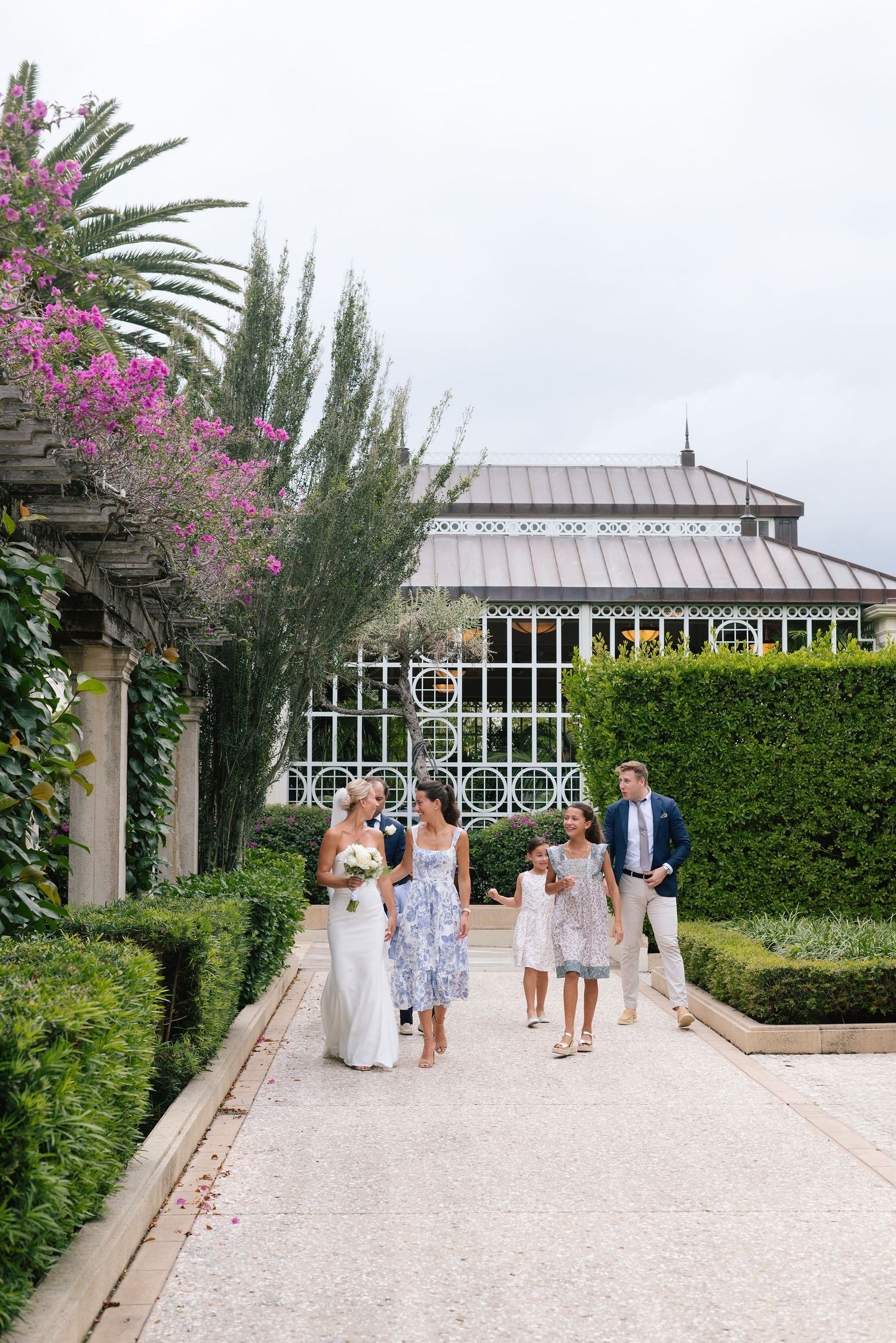 Wedding party walks along stone pathway with ornate greenhouse and flowering bushes in background.