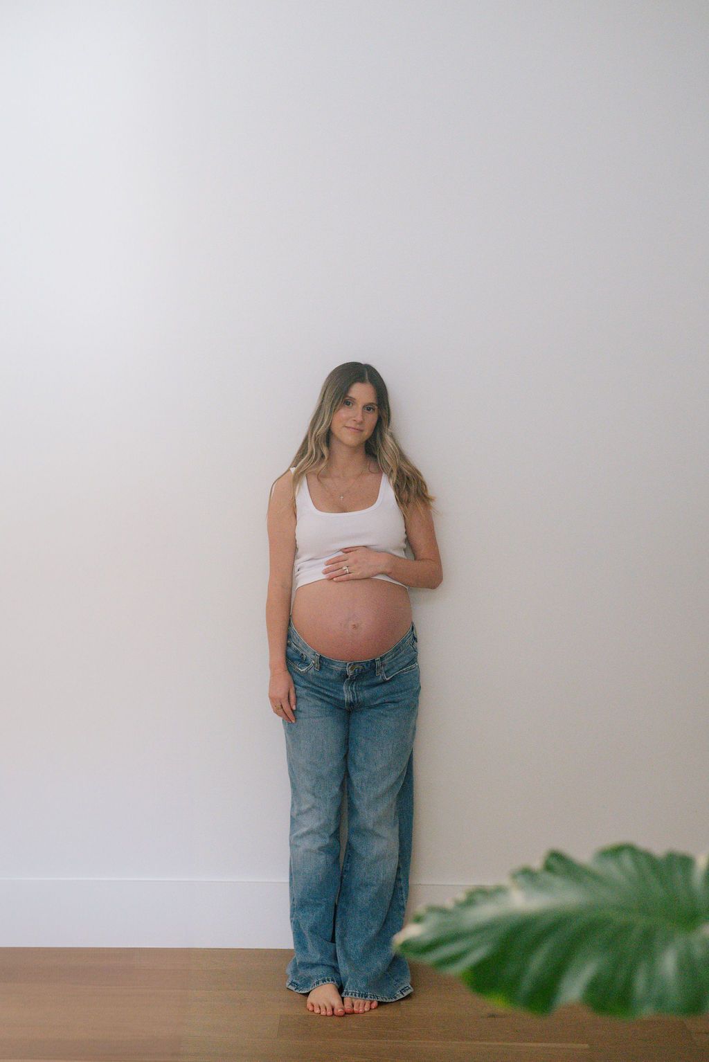 A pregnant woman in jeans and white tank top poses against a white wall.