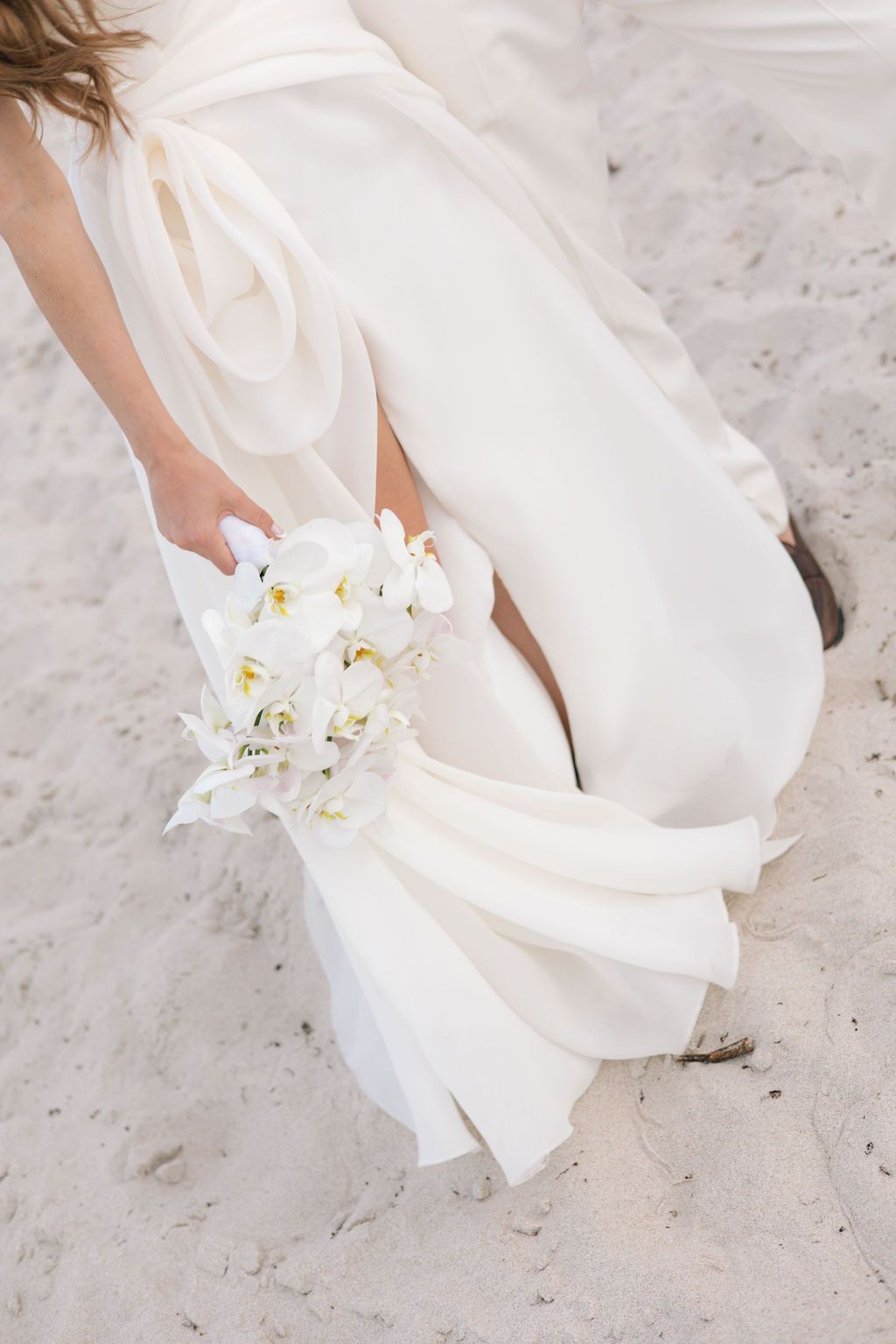 Close-up details of a white wedding dress and orchid bouquet on a beach.