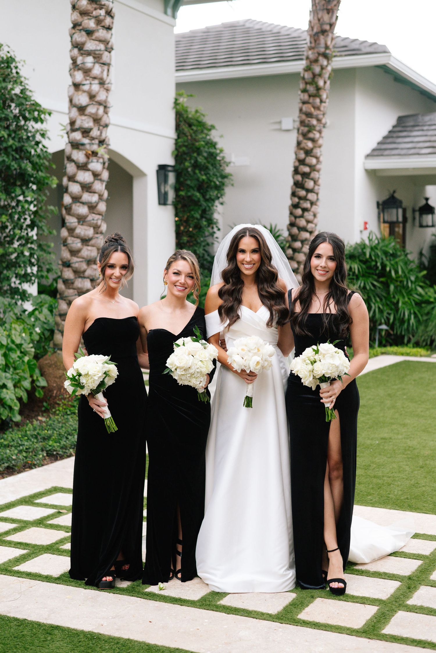 Bridal party poses outdoors in black dresses with white bouquets against a white building.