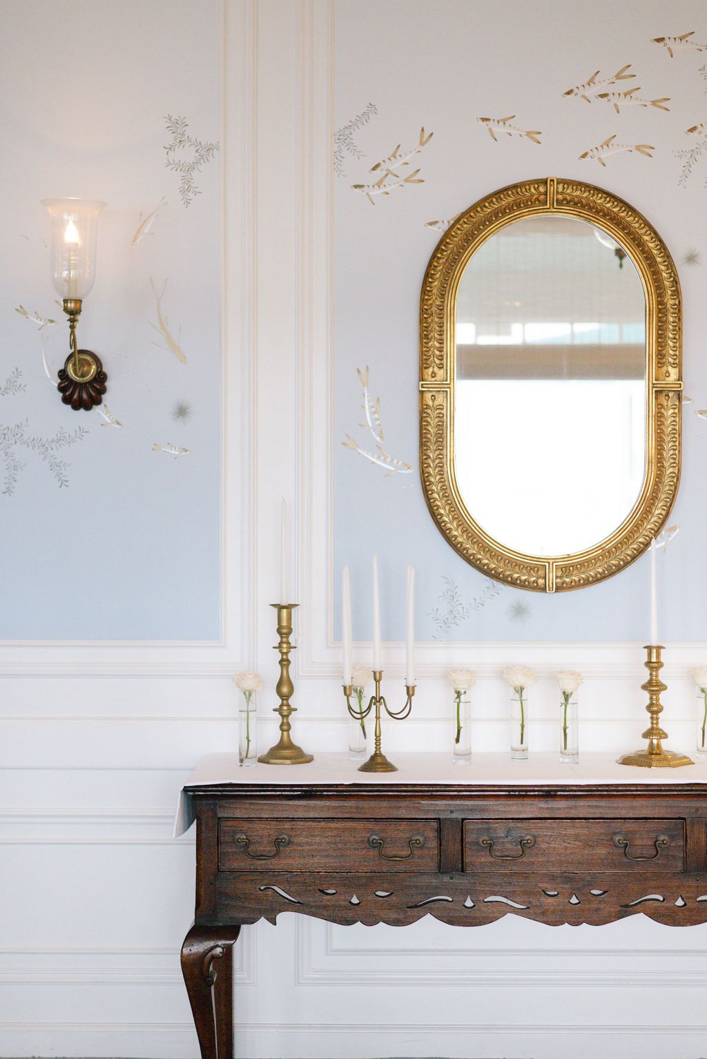 Elegant white room with gold-framed mirror and antique wooden console table displaying candelabras.