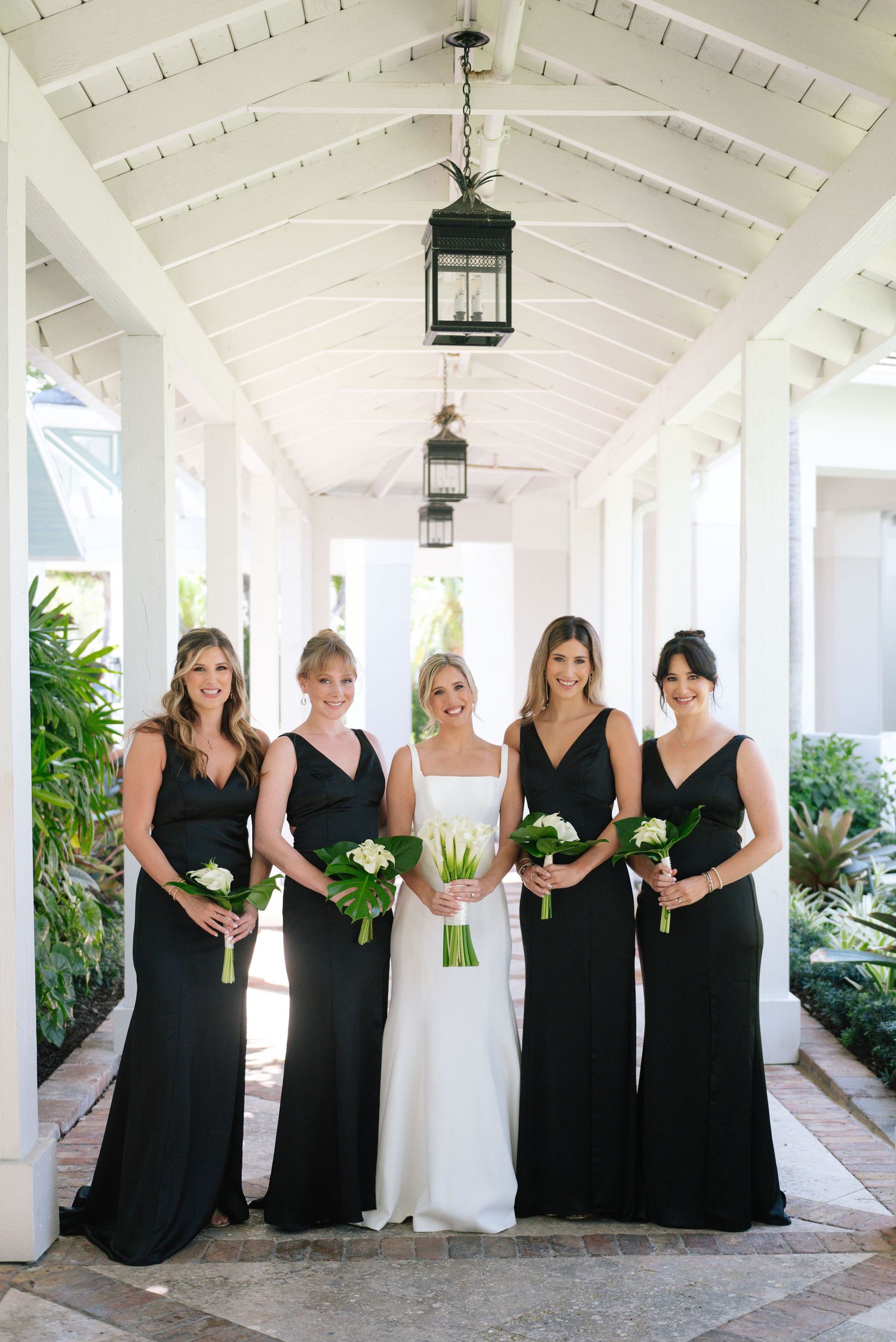 Bridesmaids in black dresses standing with bride in white under covered walkway.