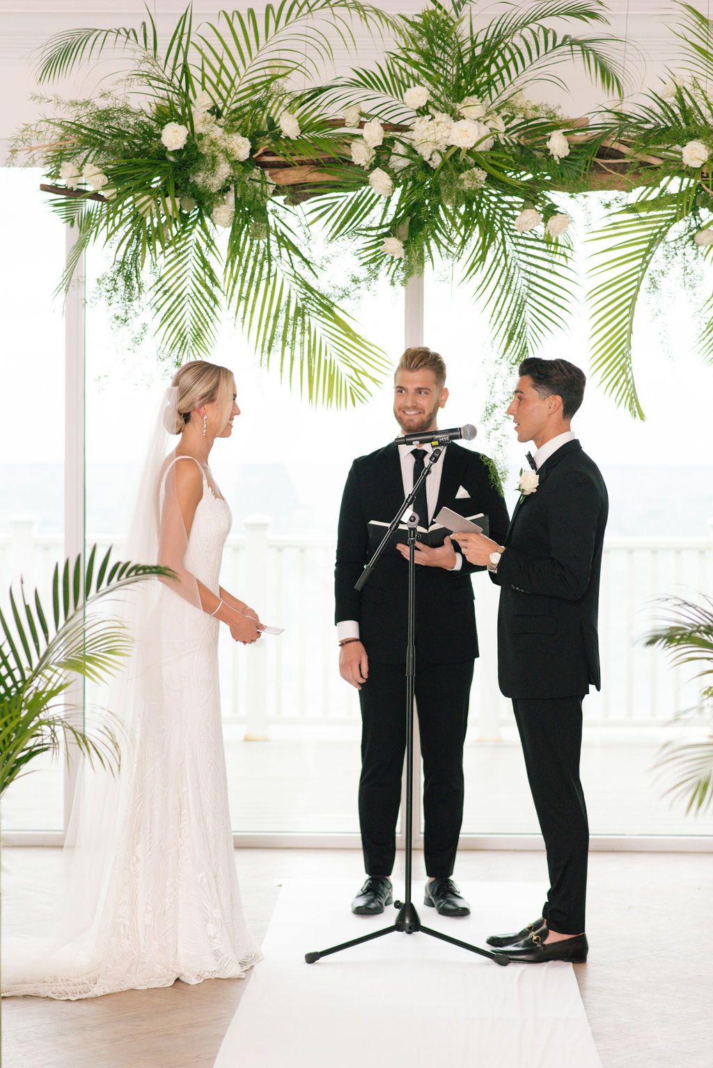 Wedding ceremony under palm leaf arch with white floral arrangements.
