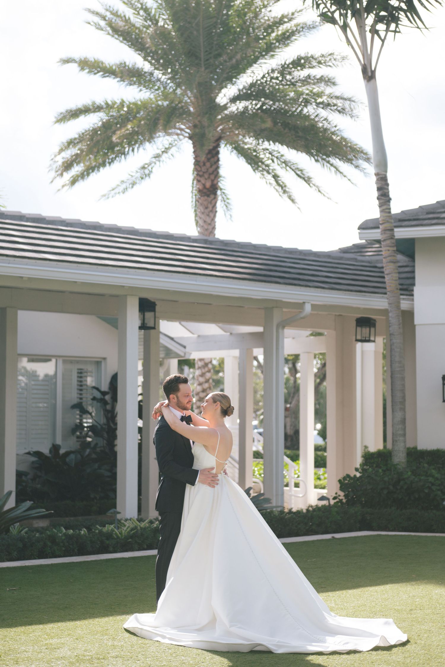 Newlyweds dancing together under palm trees outside a white columned building.