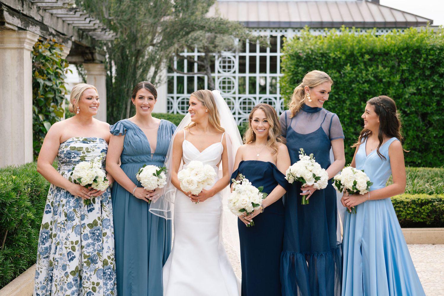 Wedding party wearing blue bridesmaid dresses and formal attire pose together in an elegant garden setting.
