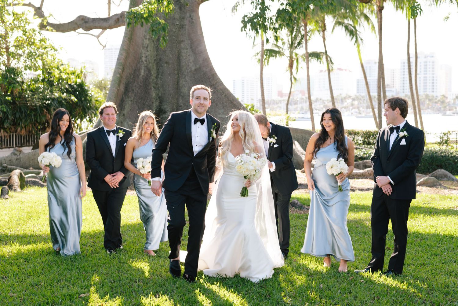 Wedding party in formal attire poses together on sunny lawn with large tree and palm trees in background.