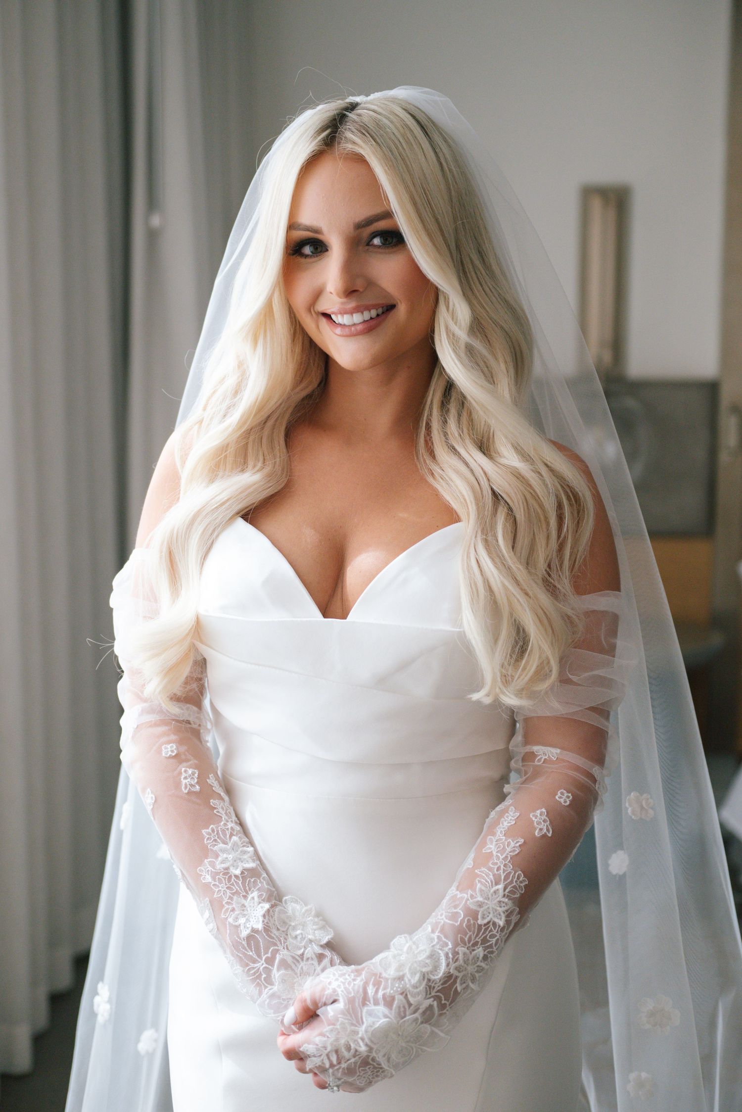 A bride wearing a classic white wedding gown with lace sleeves and a flowing veil poses indoors.