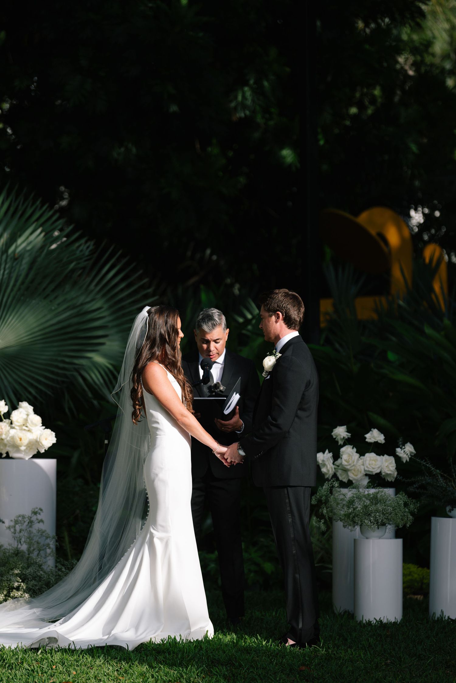 Wedding ceremony in a lush garden setting with white floral arrangements and palm fronds in the background.