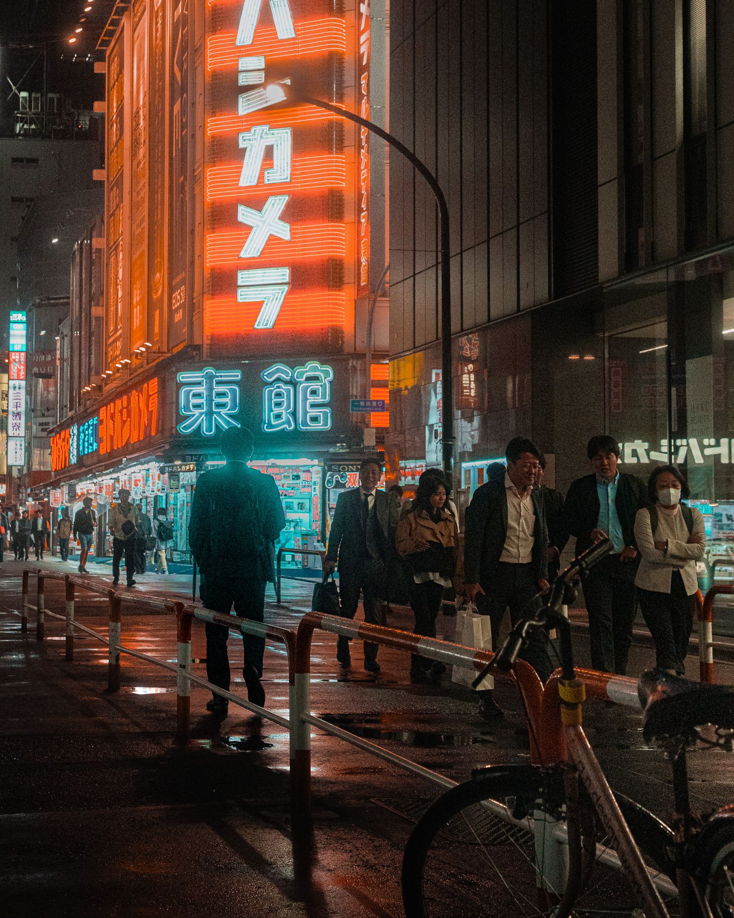 A nighttime street scene in Japan with glowing orange signs and silhouettes of people walking in the rain.