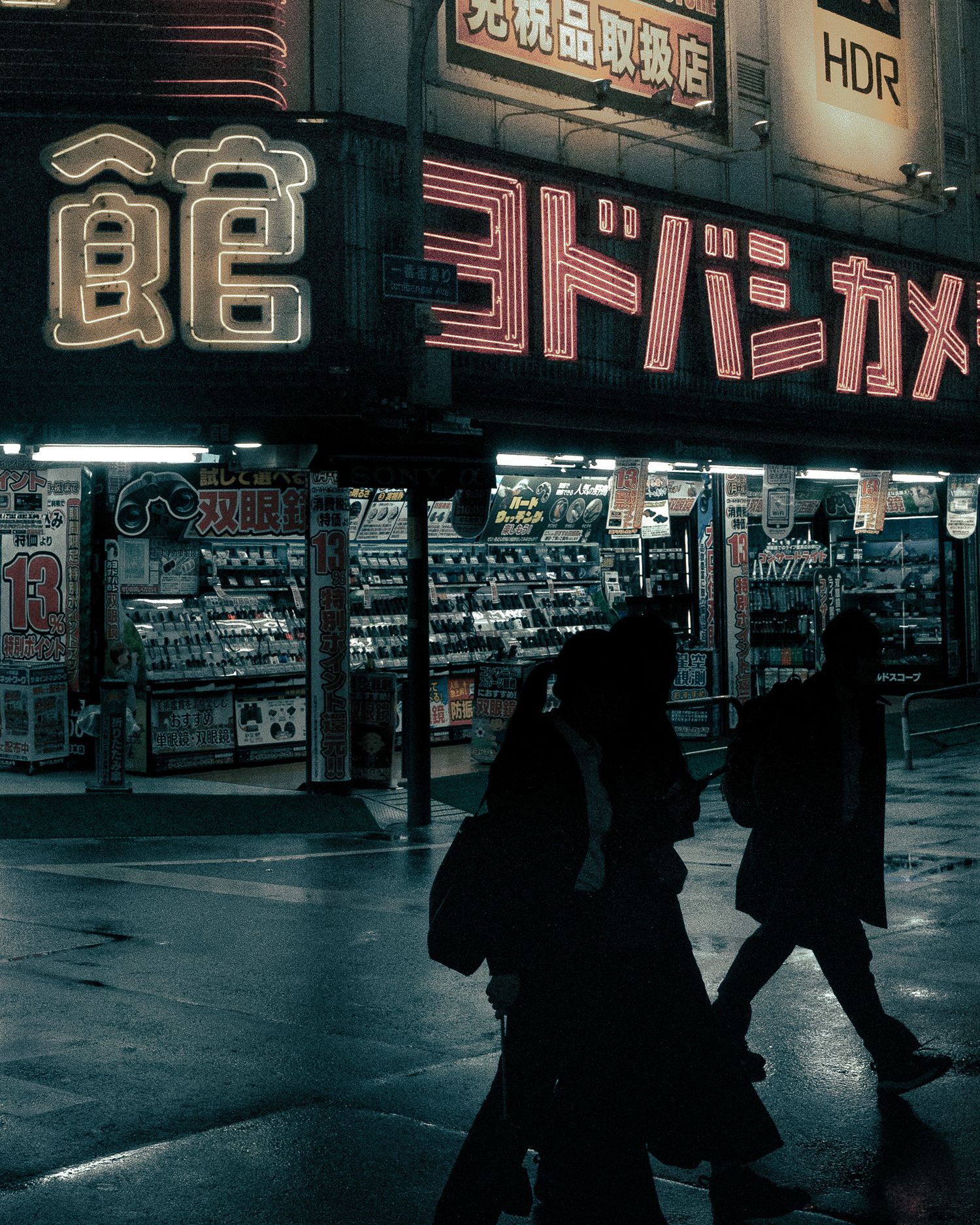 Silhouettes of pedestrians walk past neon Japanese signs and storefronts in a moody nighttime urban scene.