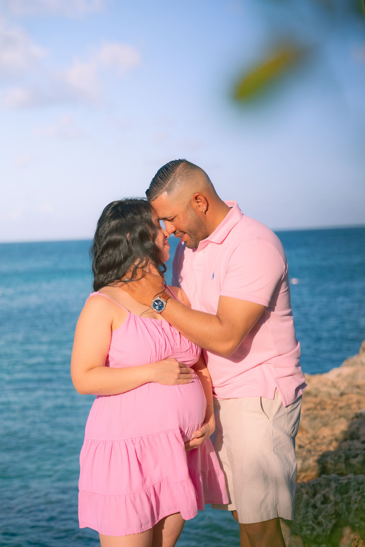 A couple shares an intimate moment during their beach maternity photoshoot in coordinating pink outfits.