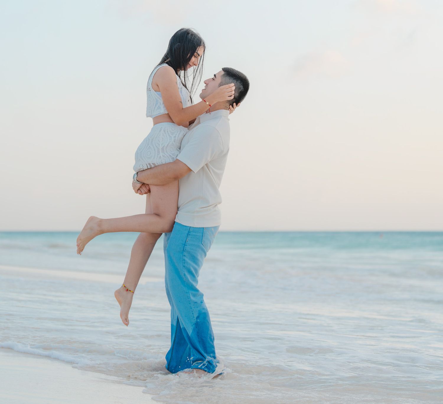 A romantic couple embraces on a serene beach at sunset, with one person lifting the other in a joyful moment.