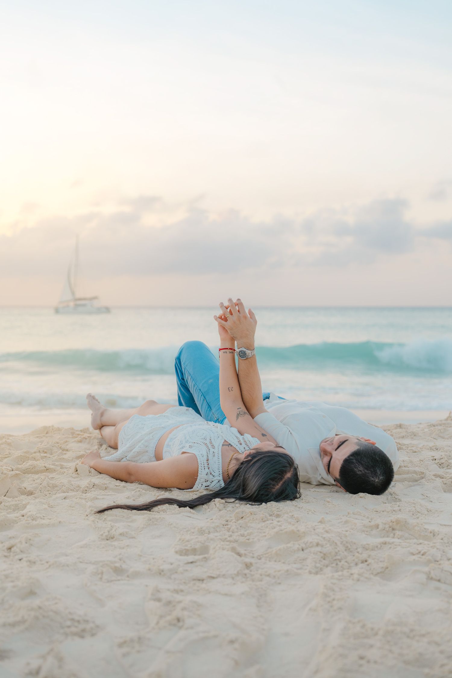 Two people lying on sandy beach holding hands with a sailboat visible in the distance.