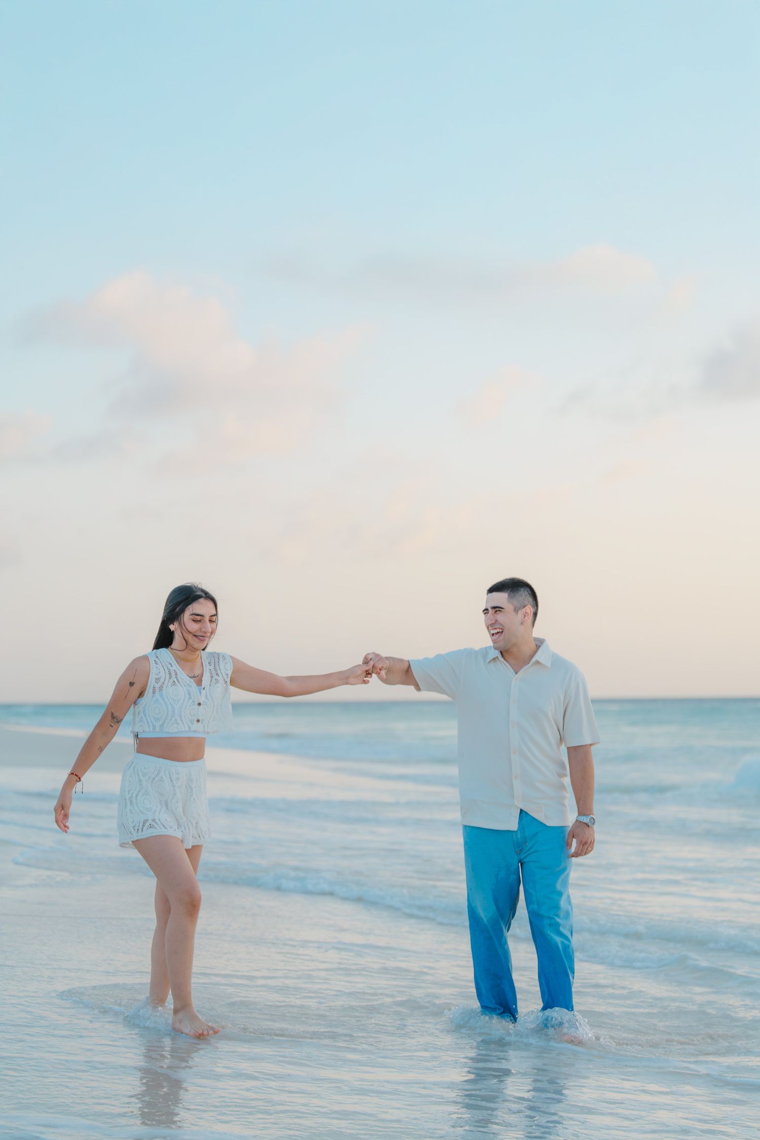 A couple holding hands while walking in shallow ocean water during sunset.