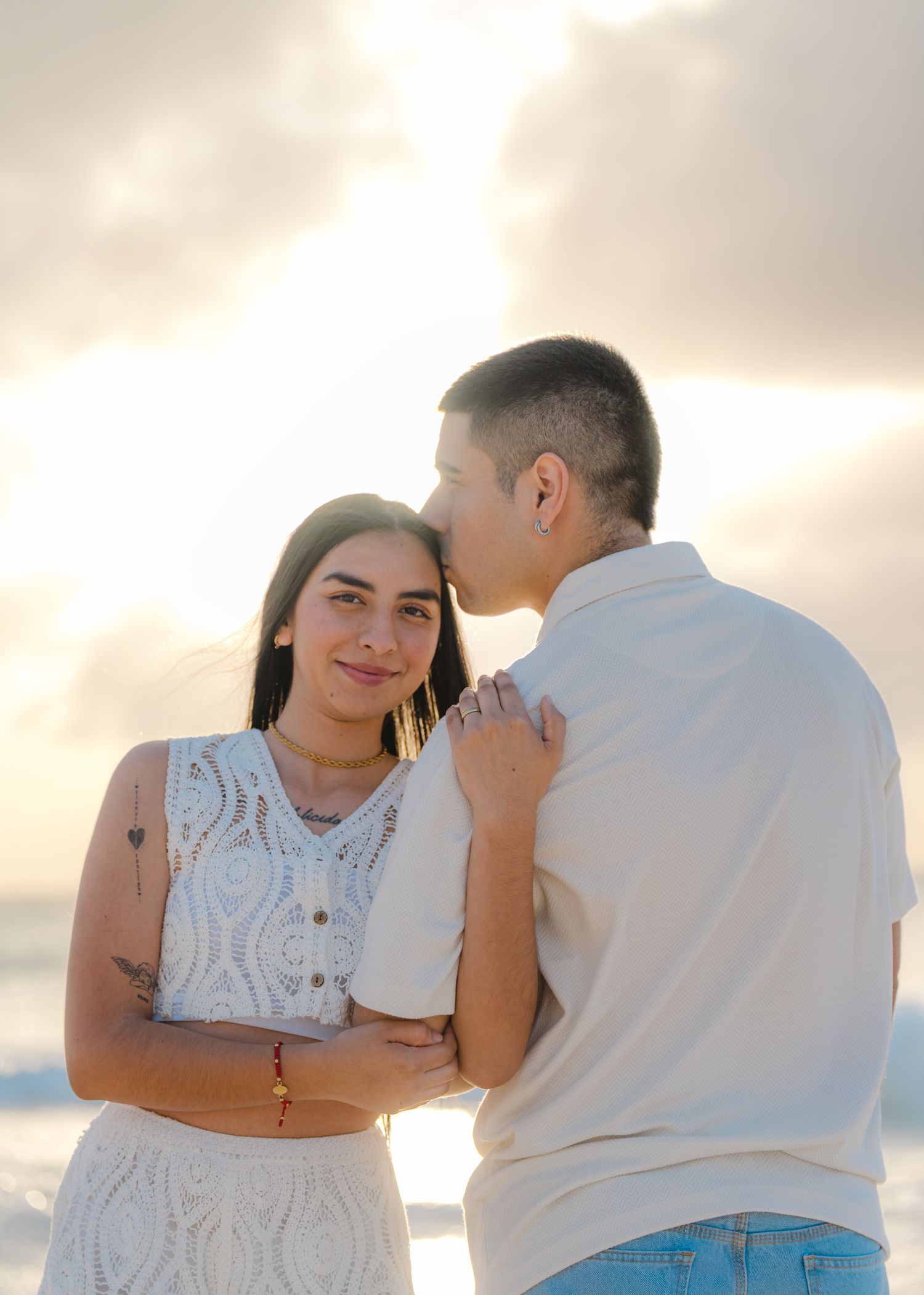 A couple in white clothing sharing an intimate moment against a sunset beach backdrop.