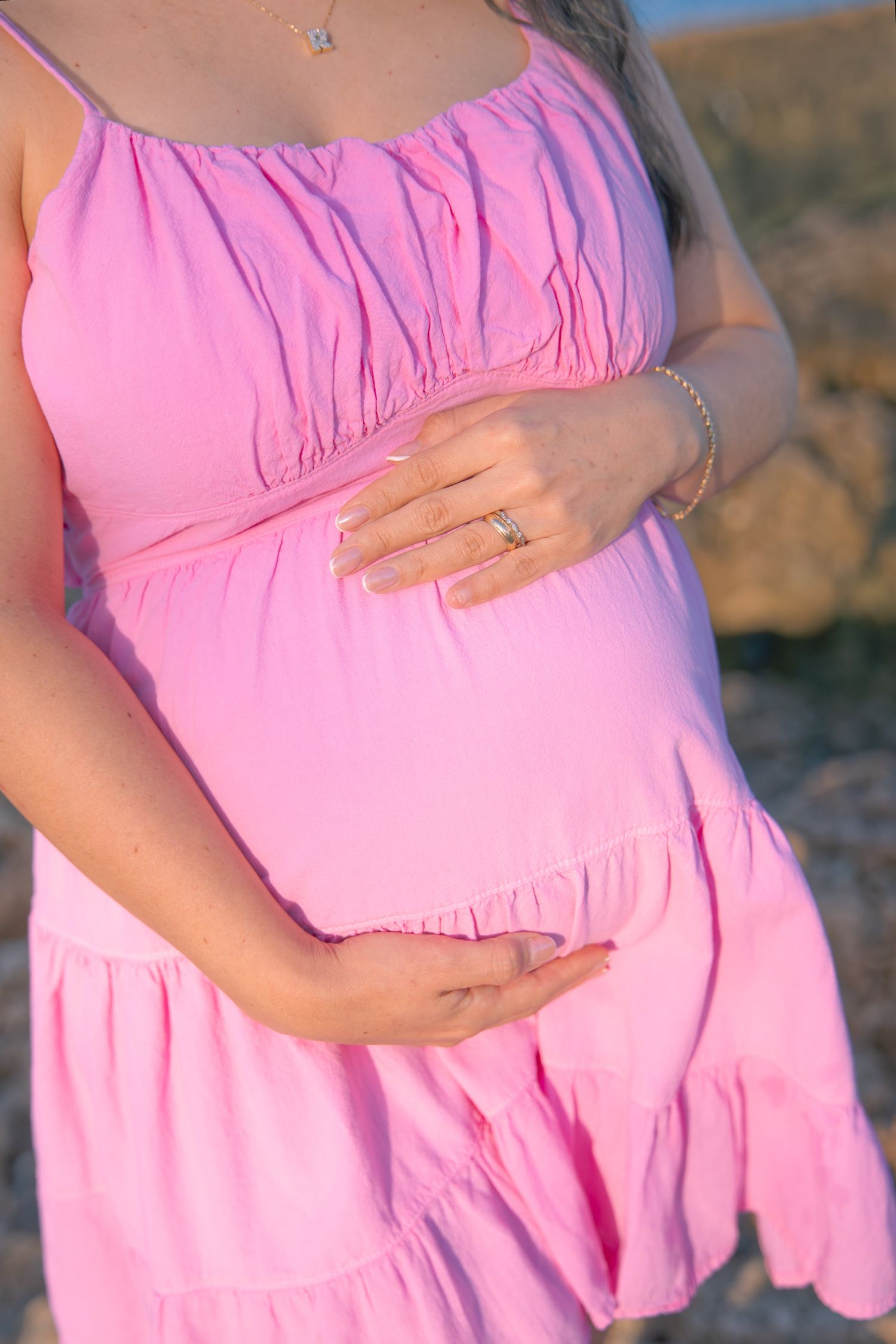 Close-up of a pregnant belly in a pink ruched maternity dress with hands gently resting on top.