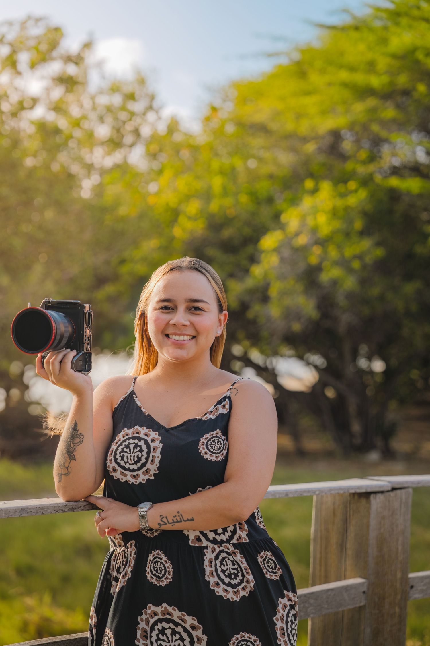 Photographer in black dress smiling while holding camera on wooden boardwalk.