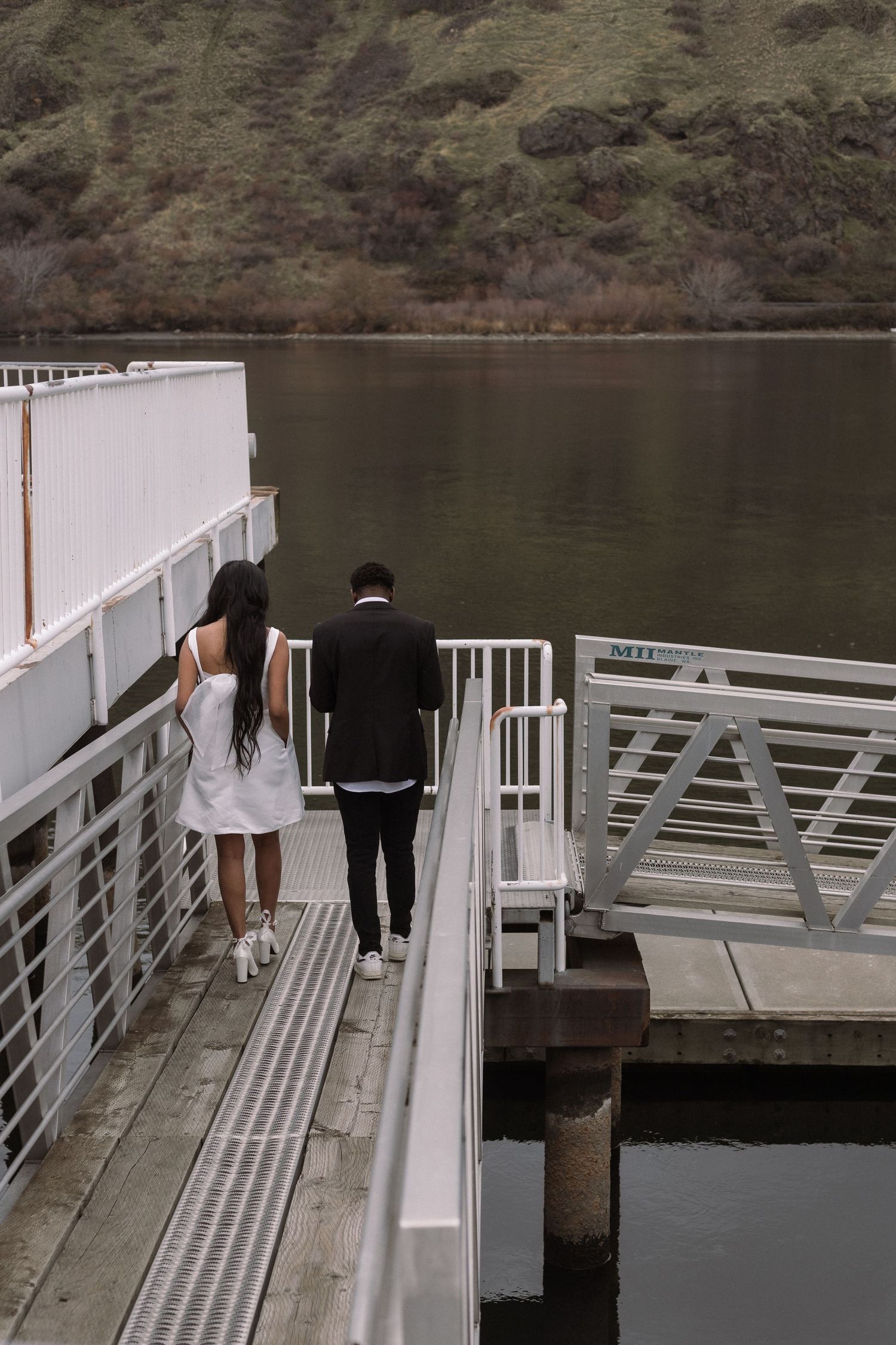 A couple walks together on a white dock overlooking a serene lake surrounded by mountains.