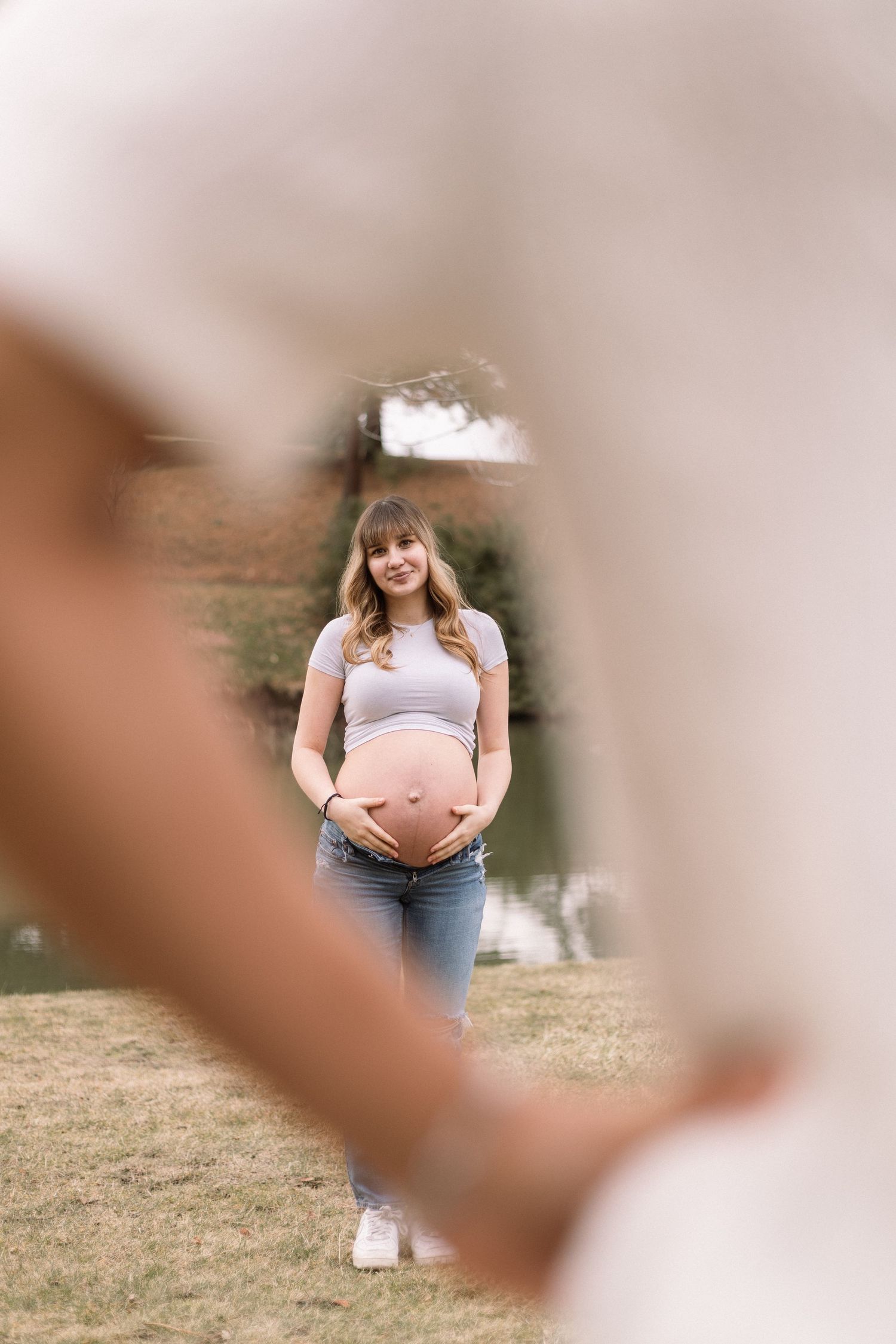 A pregnant person in a white tank top and jeans stands outdoors in a natural setting during a maternity photoshoot.