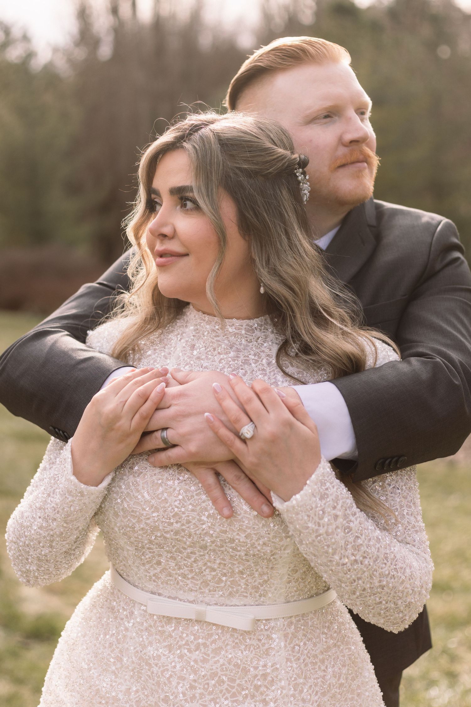 Newlywed couple embraces outdoors, bride in sparkly long sleeve dress and groom in dark suit.