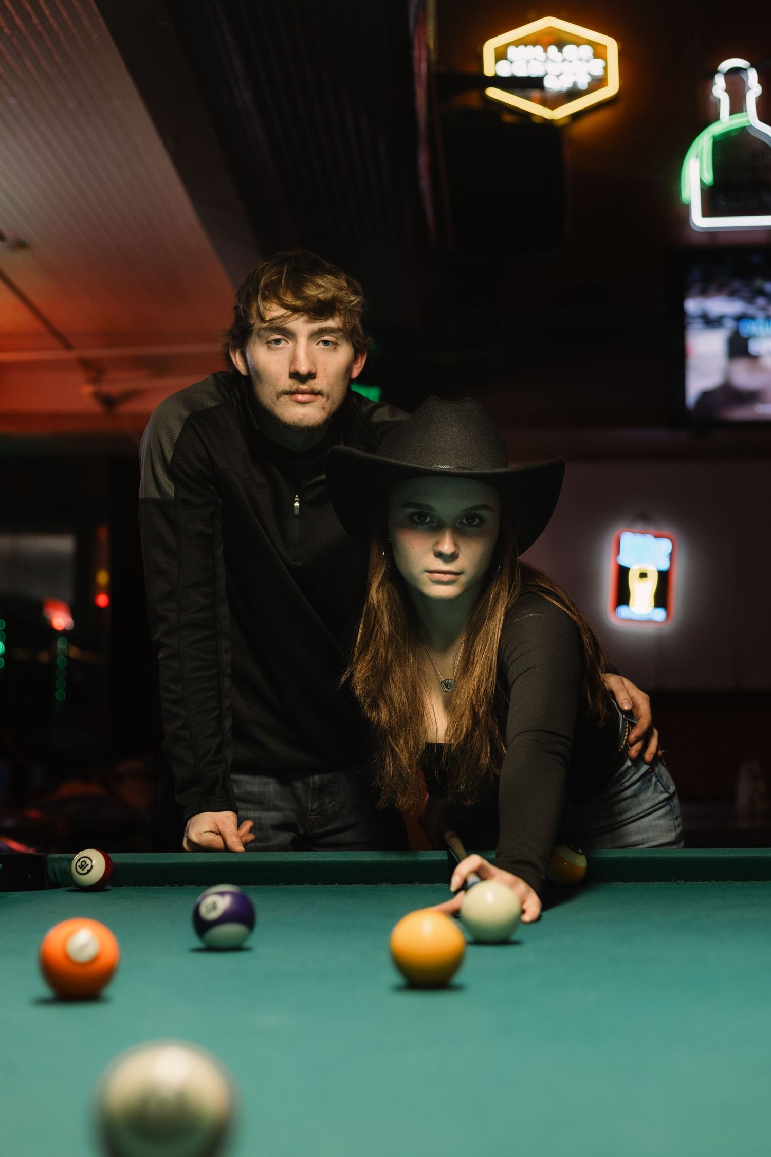 Two people playing pool in a dimly lit bar with neon signs visible in the background.