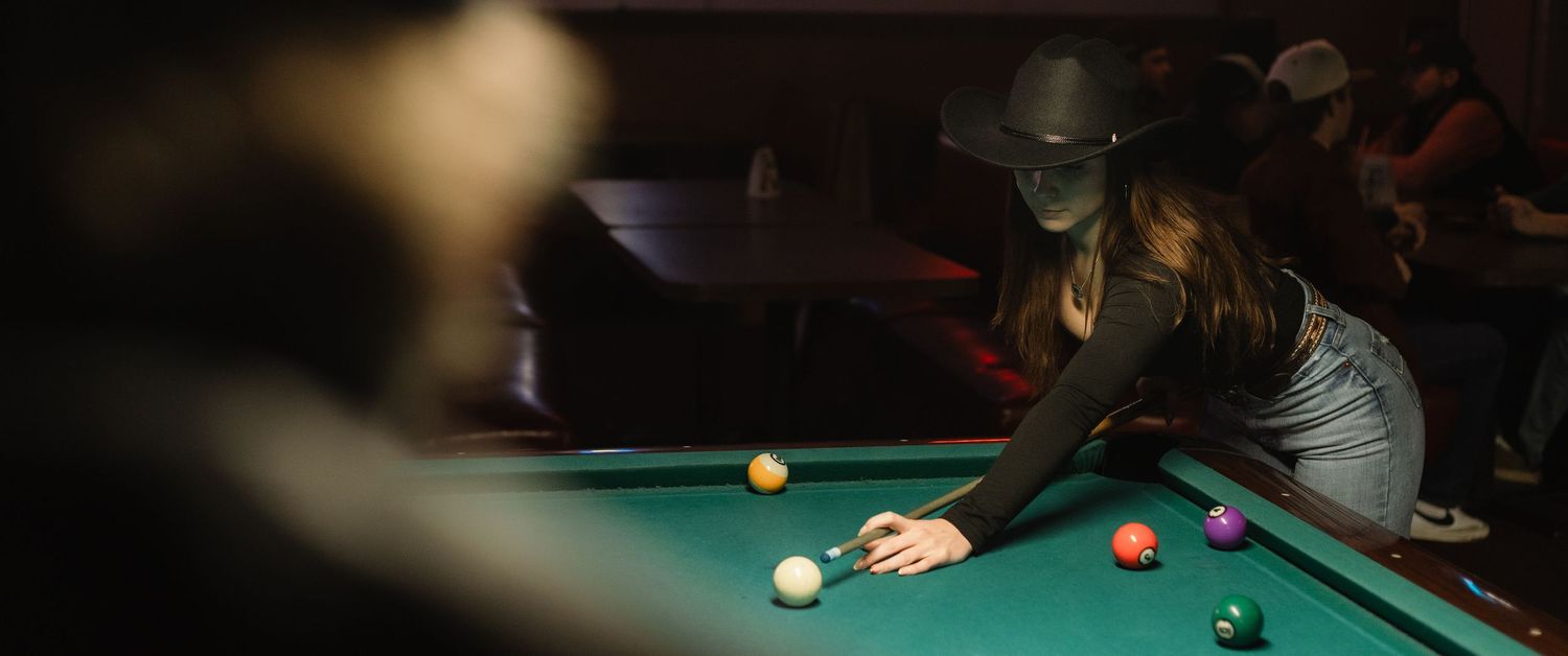 Person leaning over pool table taking aim at billiard ball in dimly lit room.