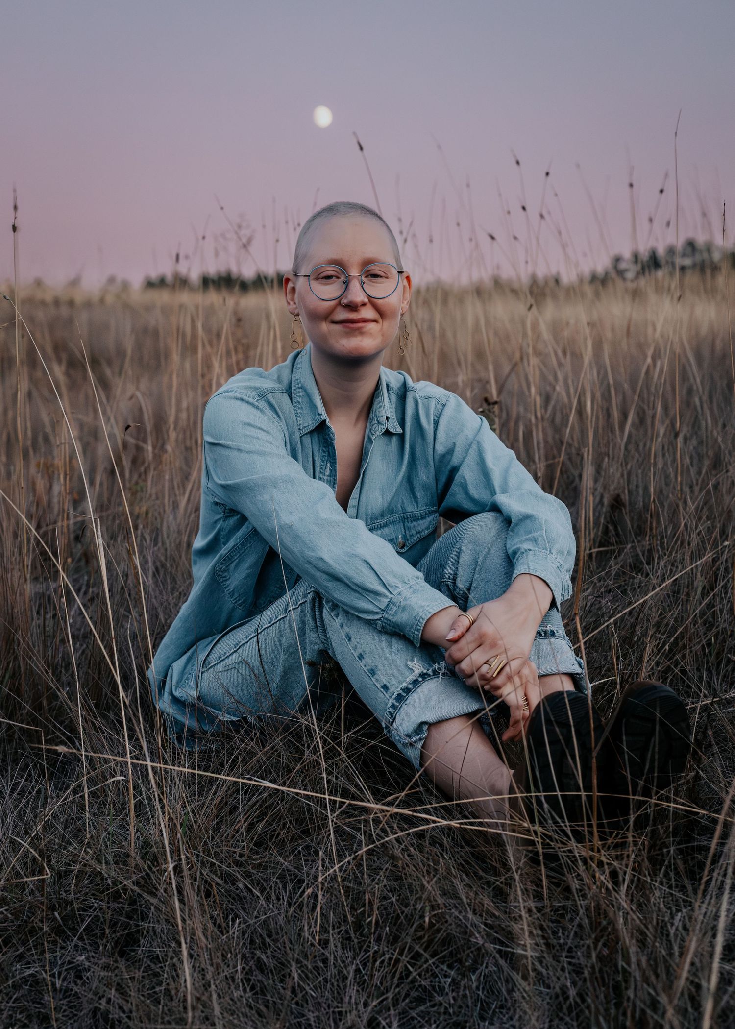 A person in denim clothing sits in a field of tall grass at dusk with the moon visible in the purple sky.