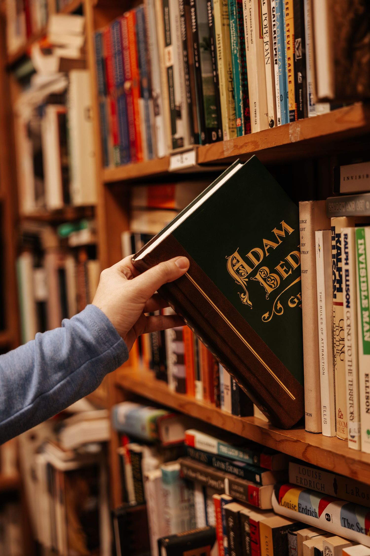 Hand reaching for a book on a wooden library shelf filled with various hardcover and paperback volumes.