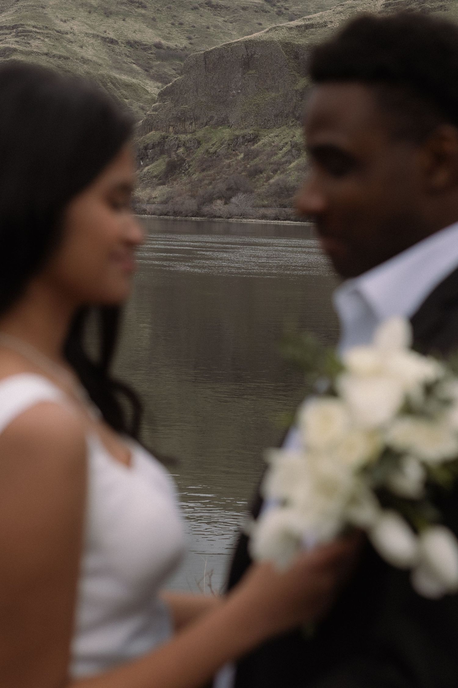 A couple in wedding attire poses by a scenic lake with mountains in the background.