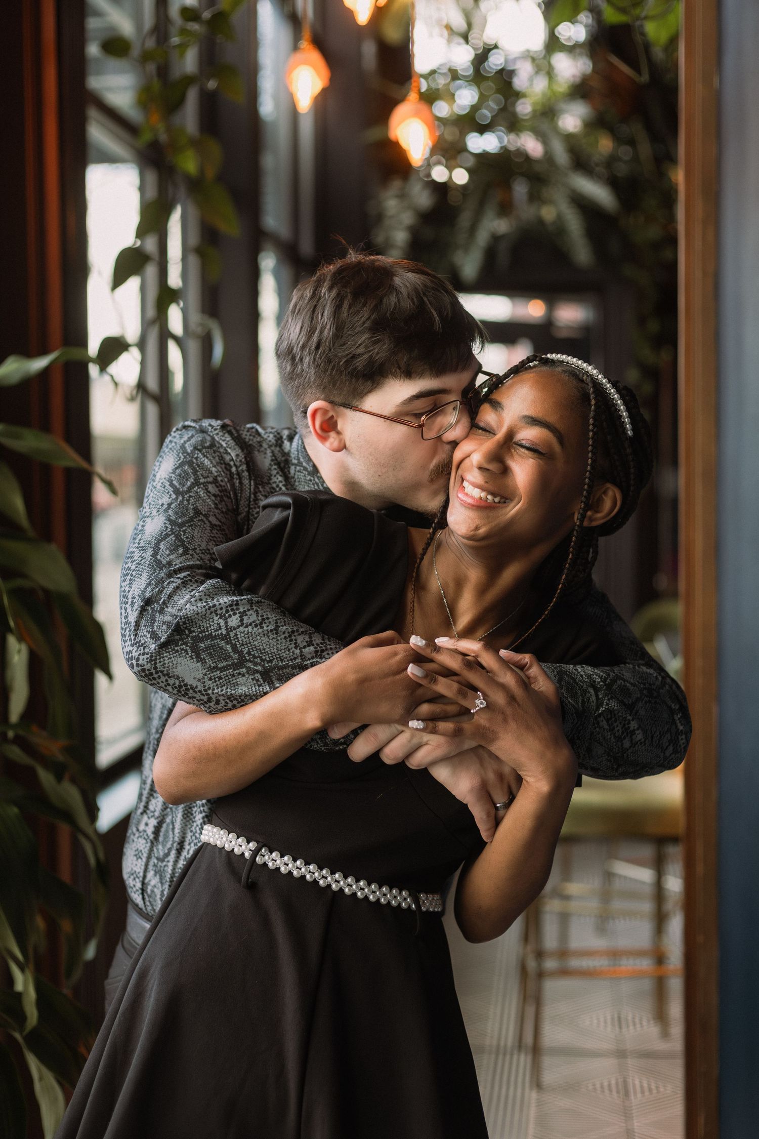 A couple embracing and kissing in a cozy, dimly lit setting with plants and decor in the background.