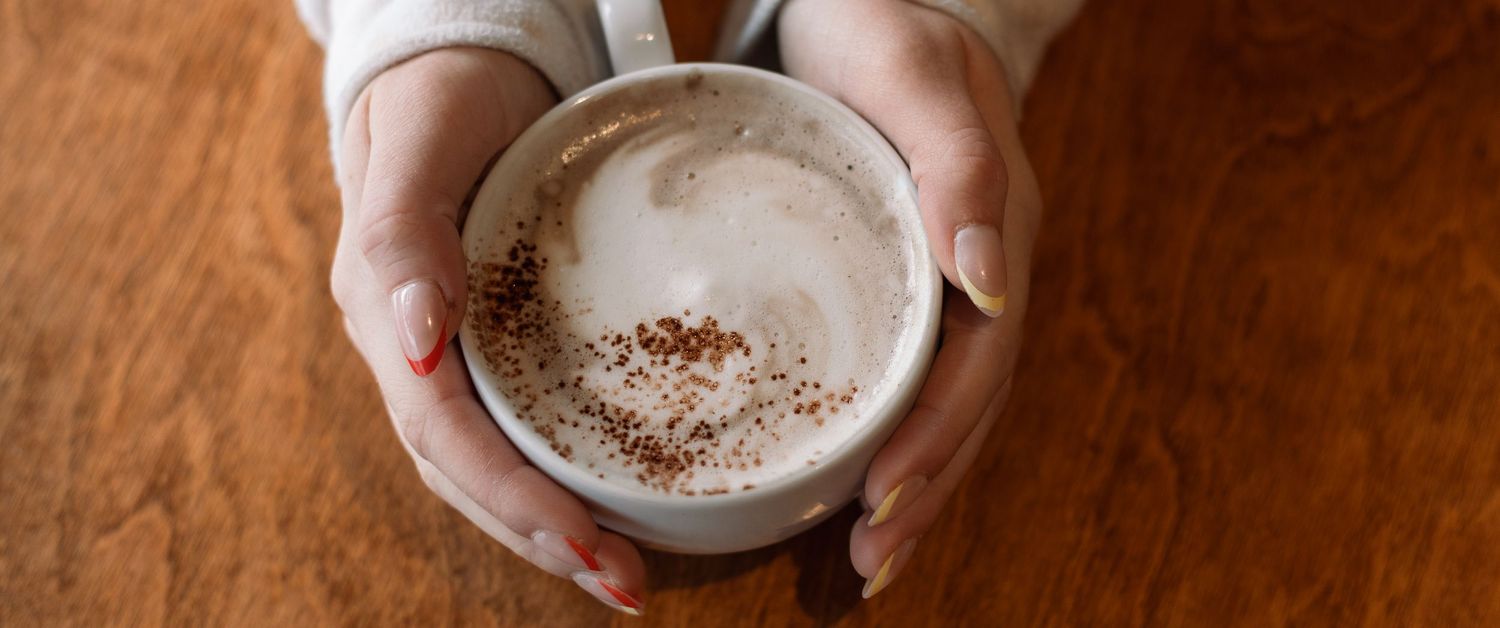Close-up of hands holding white ceramic cup with frothy coffee drink.