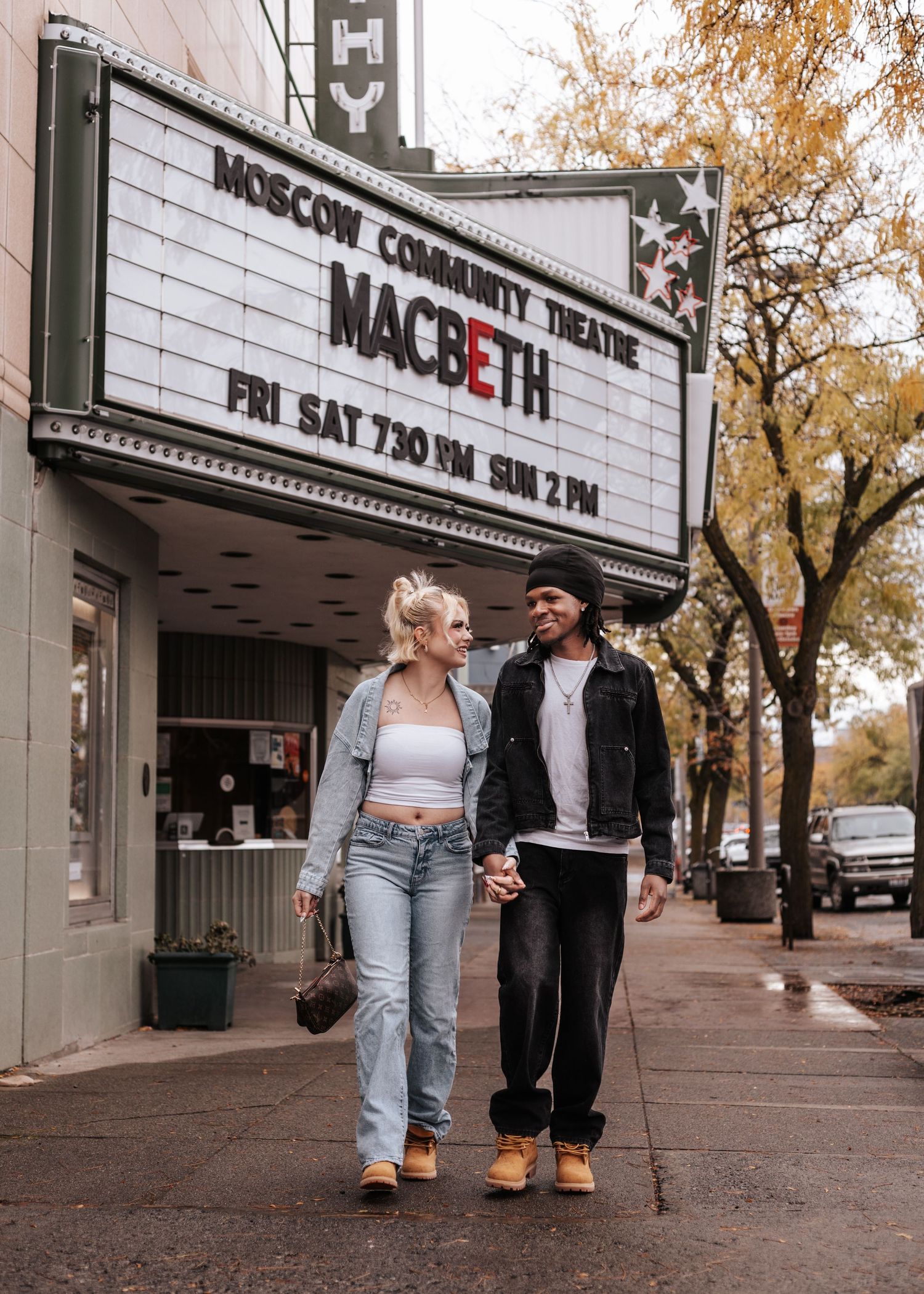 A couple walks together outside a theater marquee showing Macbeth, holding hands on an autumn day.